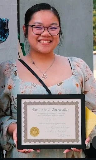Medium shot of Melayla standing with her certificate wearing a blue top with floral details.