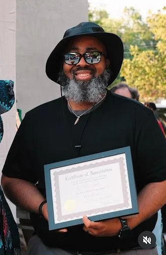 Medium shot of Mr. Baxter standing center frame wearing a black T-shirt, sunglasses, and a black bucket hat. He he holding the certificate of his award.