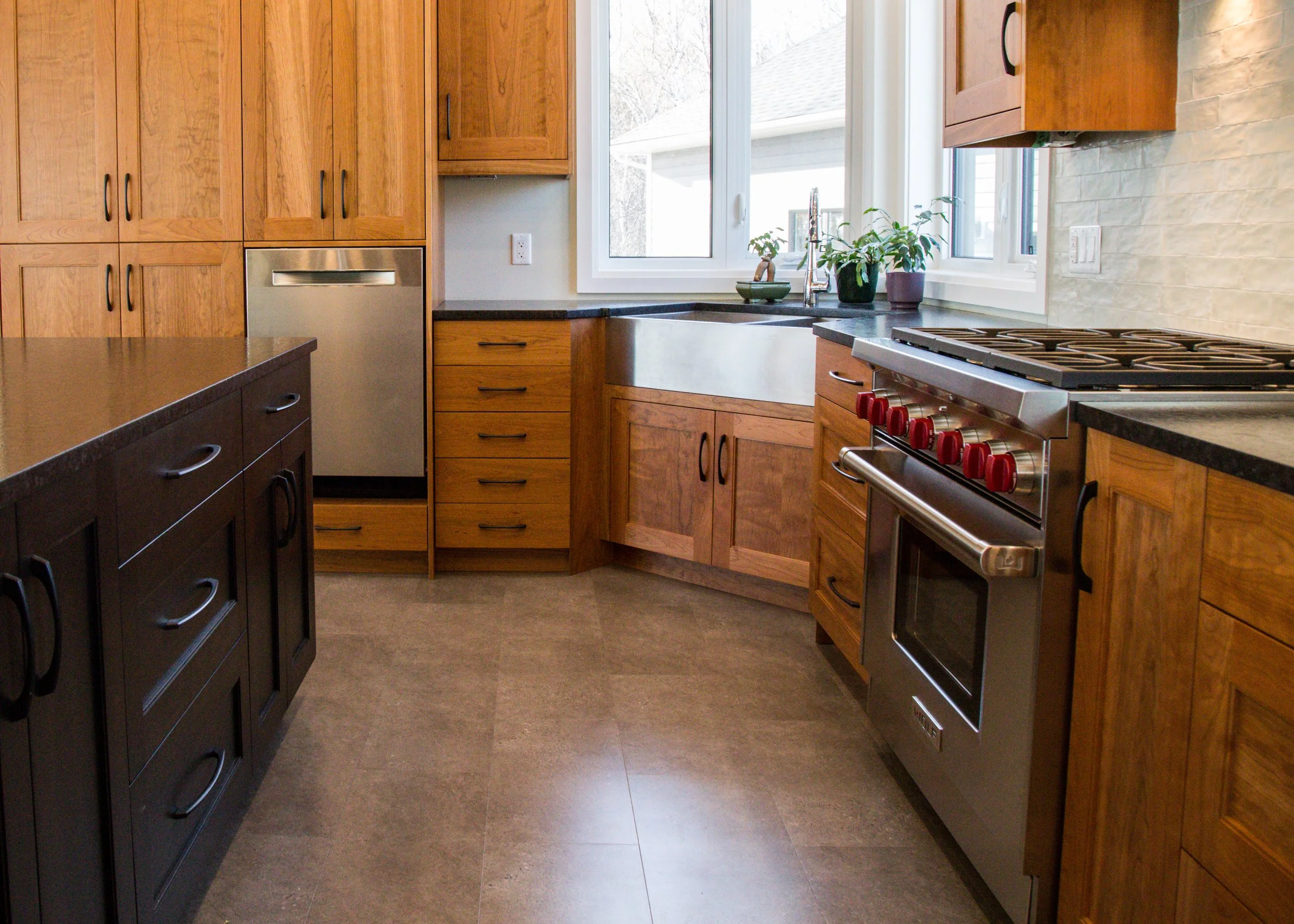 A modern kitchen with wooden cabinets, black countertops, a stainless steel oven with red knobs, a large farmhouse sink below windows, and potted plants on the windowsill.