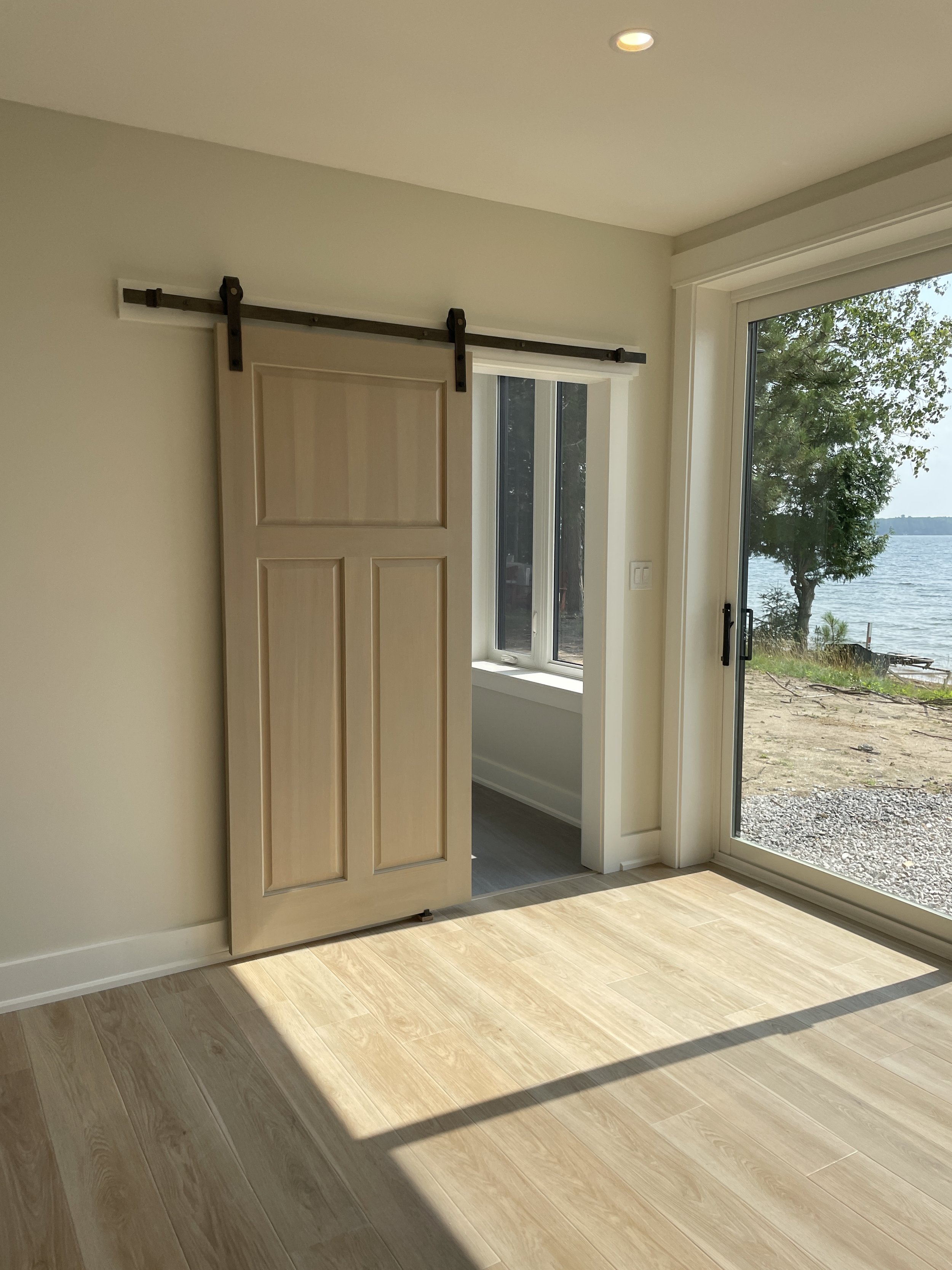 Empty room with a sliding glass door and a barn door leading to a sunroom, overlooking a body of water with greenery outside.