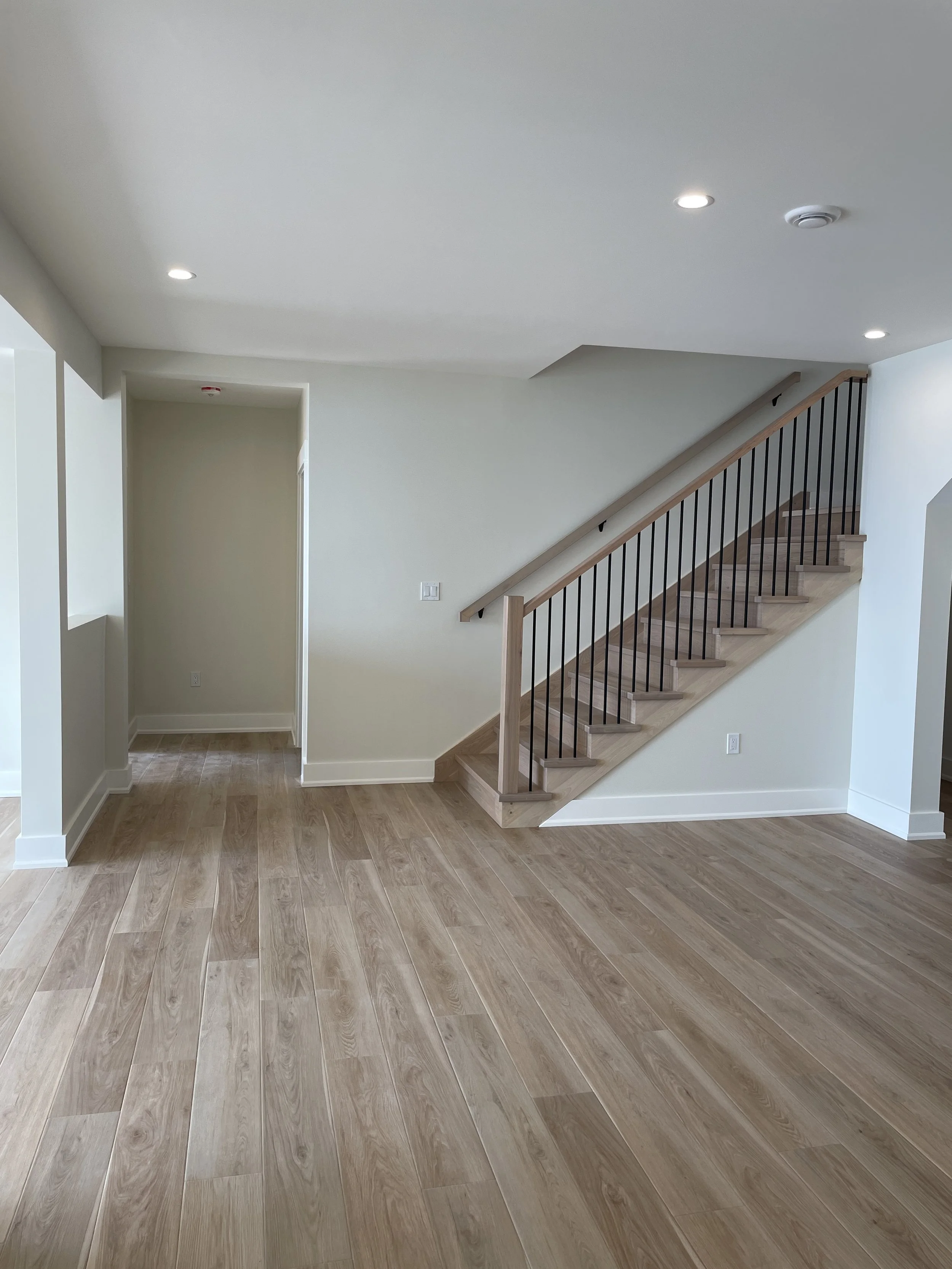 Empty room with light wood flooring, beige walls, staircase with wooden steps and black metal balusters leading upstairs, recessed lighting in the ceiling.
