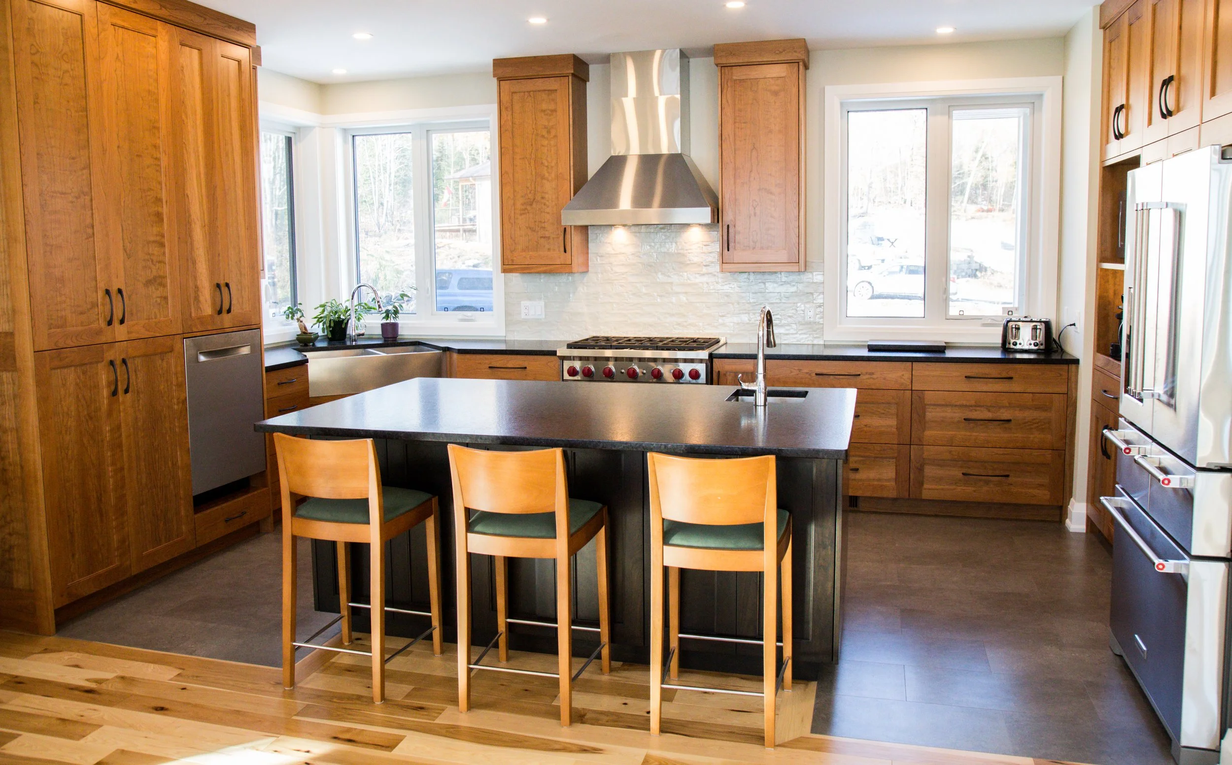 Kitchen with wooden cabinets, black countertops, a central island with three wooden chairs, stainless steel appliances including a range and refrigerator, large windows, and a brick backsplash.