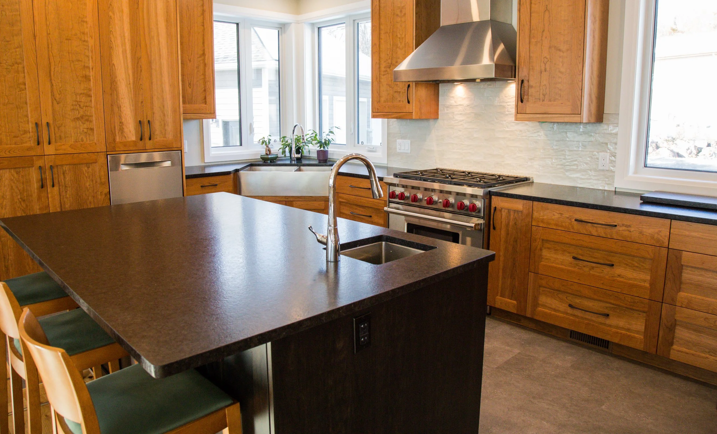 Modern kitchen with wooden cabinets, black countertops, a stainless steel stove and range hood, a kitchen island with a sink, and large windows allowing natural light.
