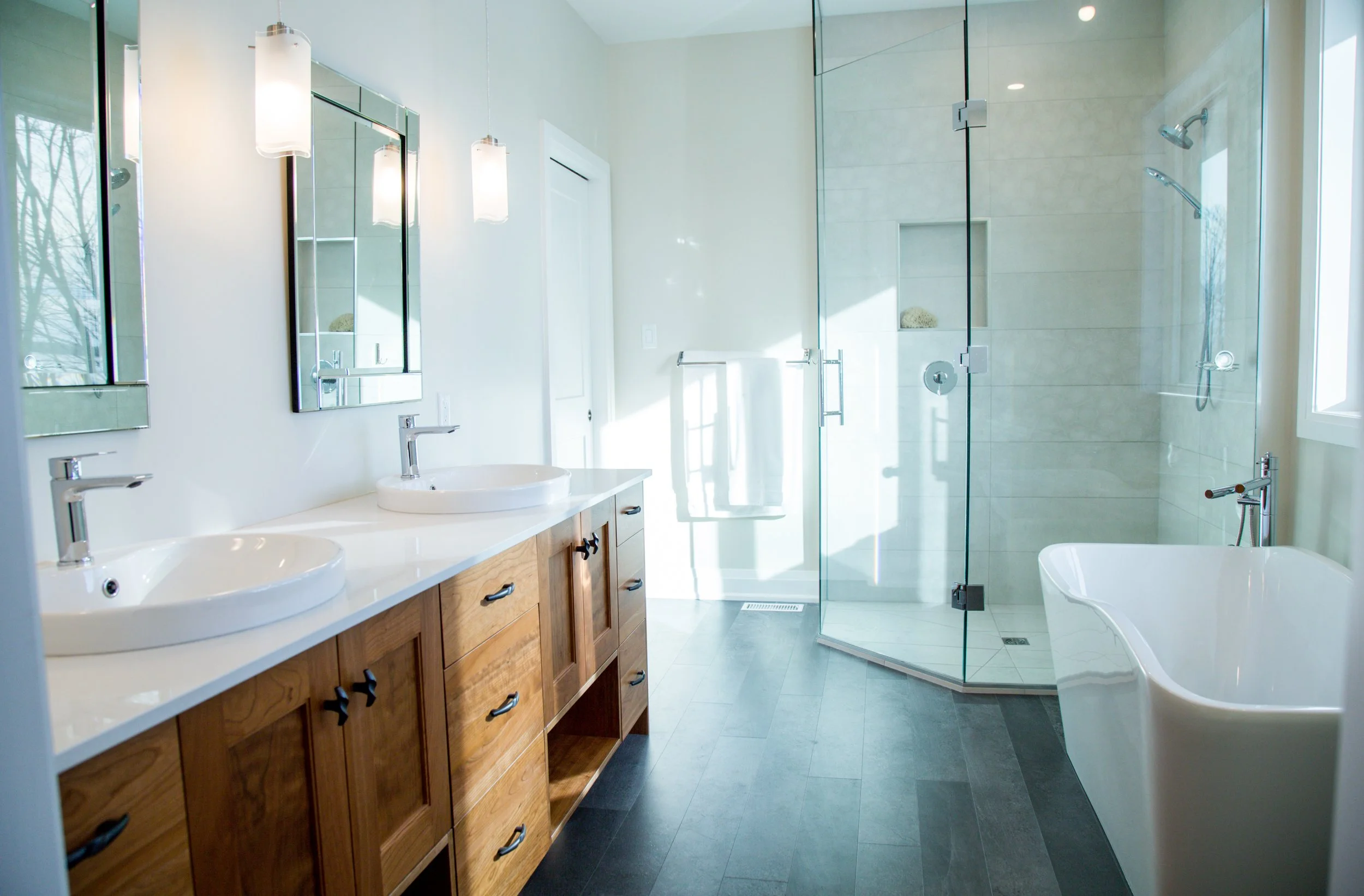 Modern bathroom with dual sinks, wooden vanity, large mirror, glass-enclosed shower, and a free-standing bathtub, with natural light coming in through a window.