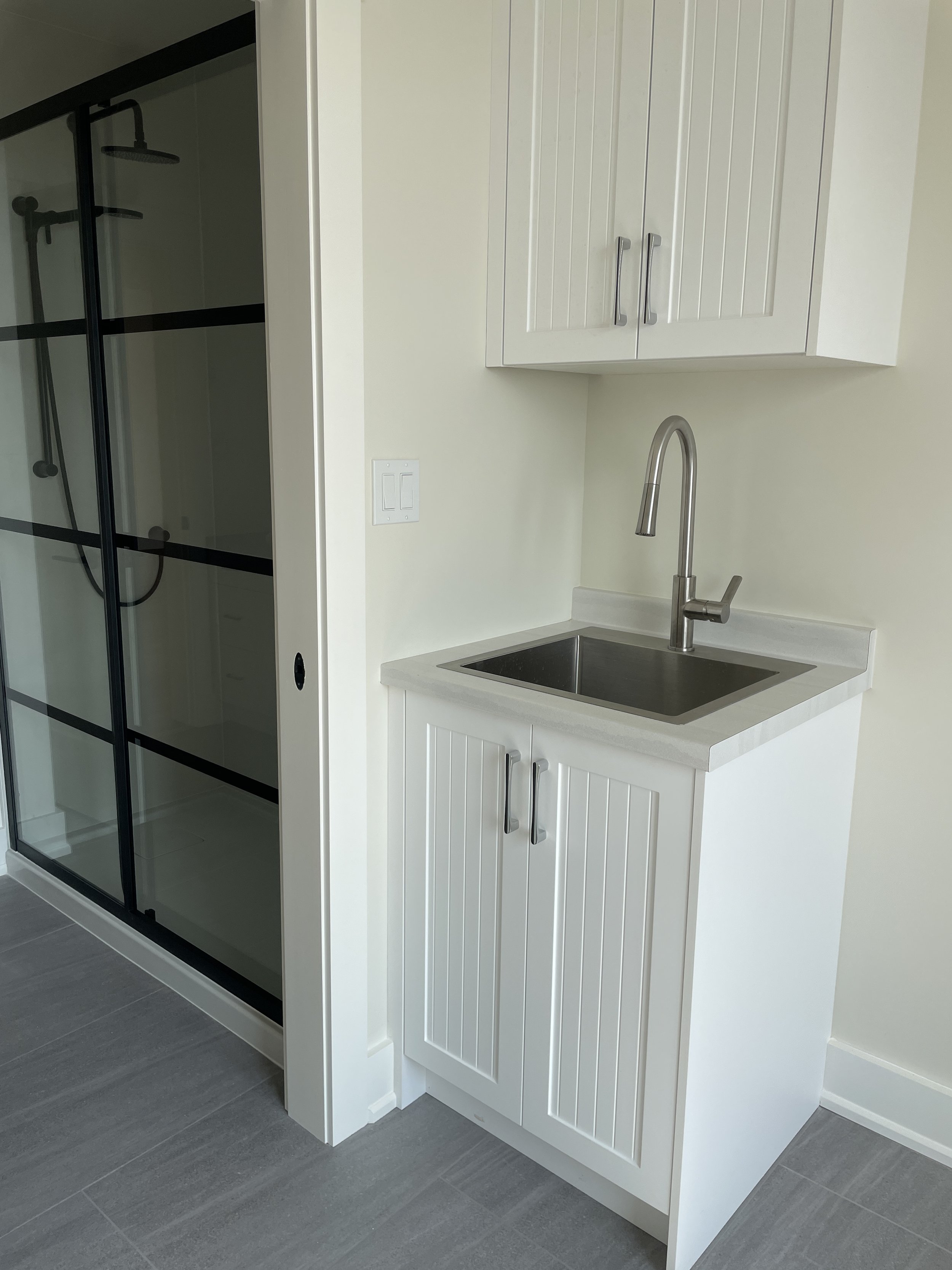 Small white sink cabinet with a stainless steel faucet and a wall-mounted cabinet above it, next to a glass-enclosed walk-in shower.