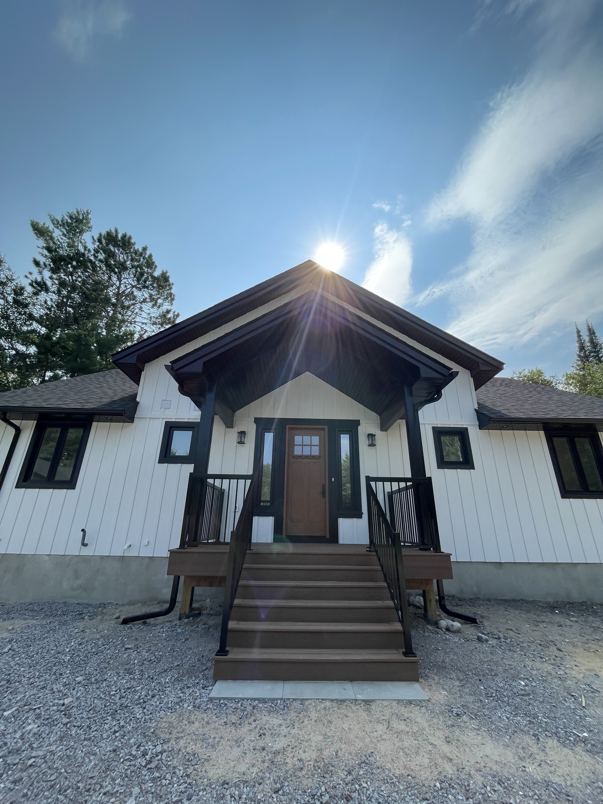 Front view of a modern house with a staircase leading to a wooden front door, white siding, black window frames, surrounded by trees, under a blue sky with sun and clouds.