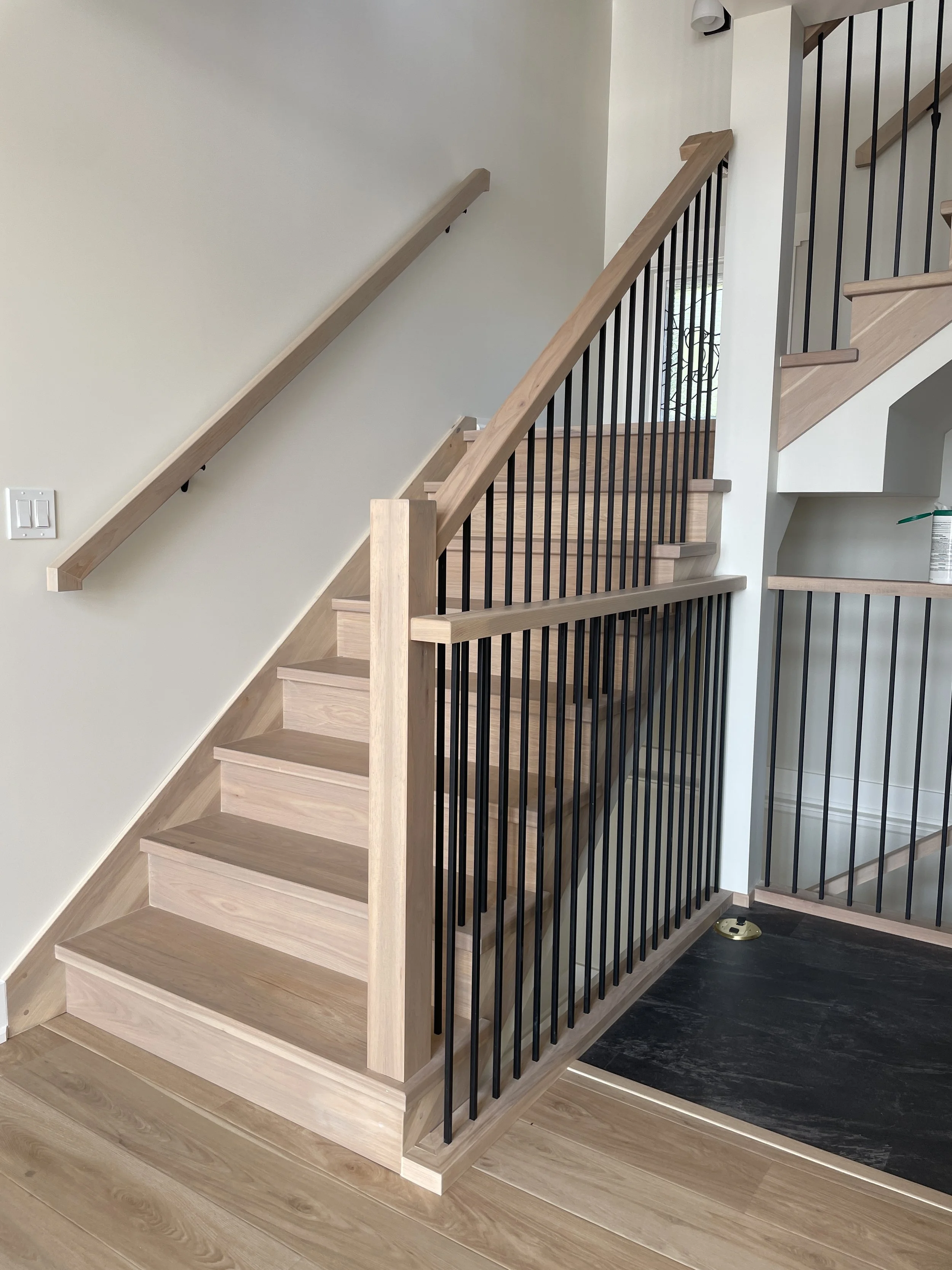 Interior view of a wooden staircase with black metal railings and a side handrail, with light-colored wooden flooring and wall.