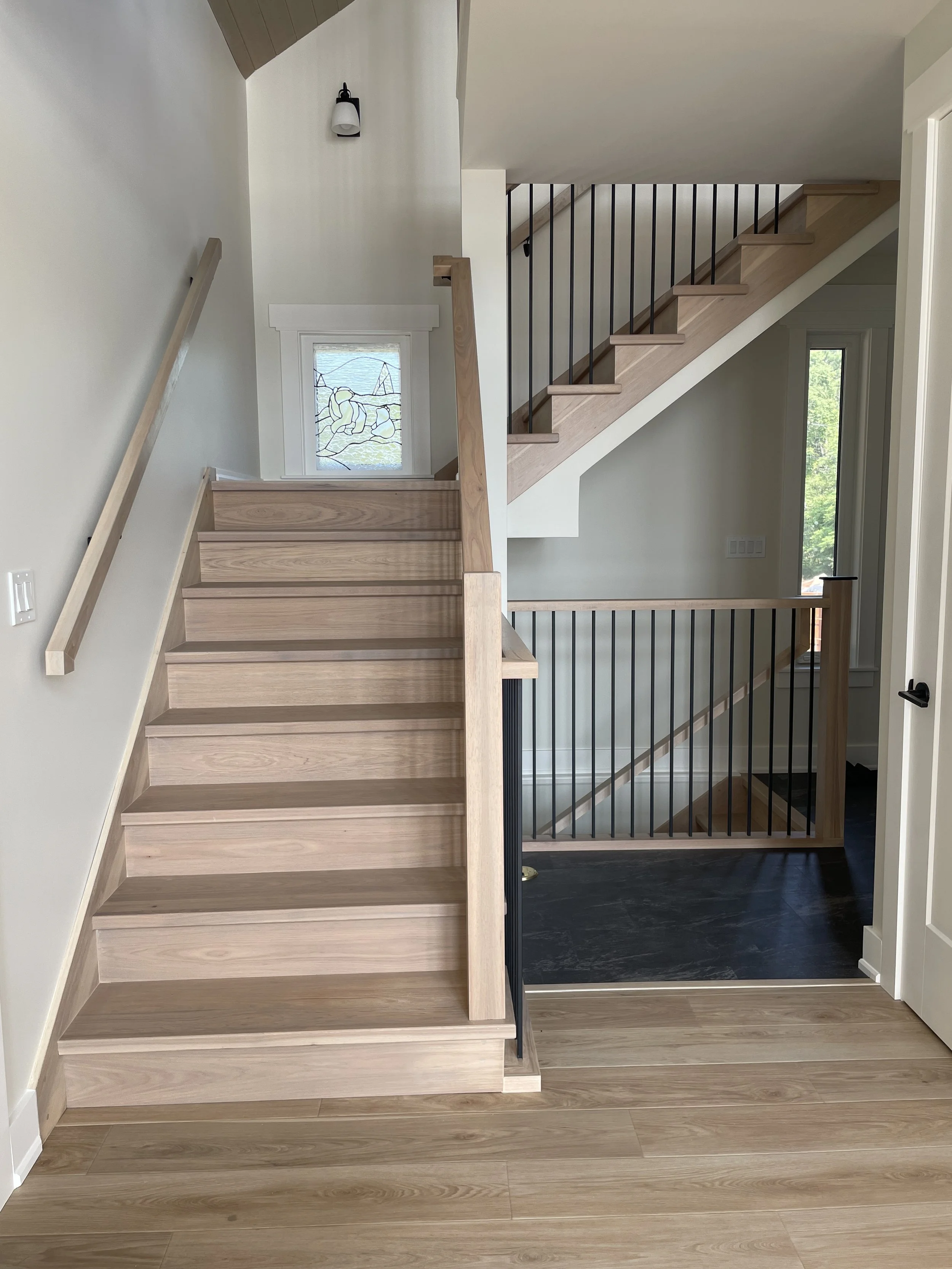 Interior staircase with light wood steps, black metal railings, and a white wall. A small window with stained glass is visible on the upper wall, and a black wall-mounted light fixture is above it.