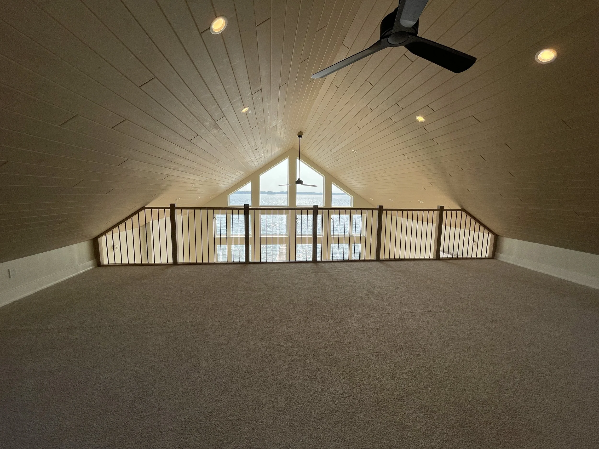 Empty attic space with sloped wood-paneled ceiling, beige carpeted floor, and a large windowed wall providing a view of water and sky, with ceiling fan and recessed lights.
