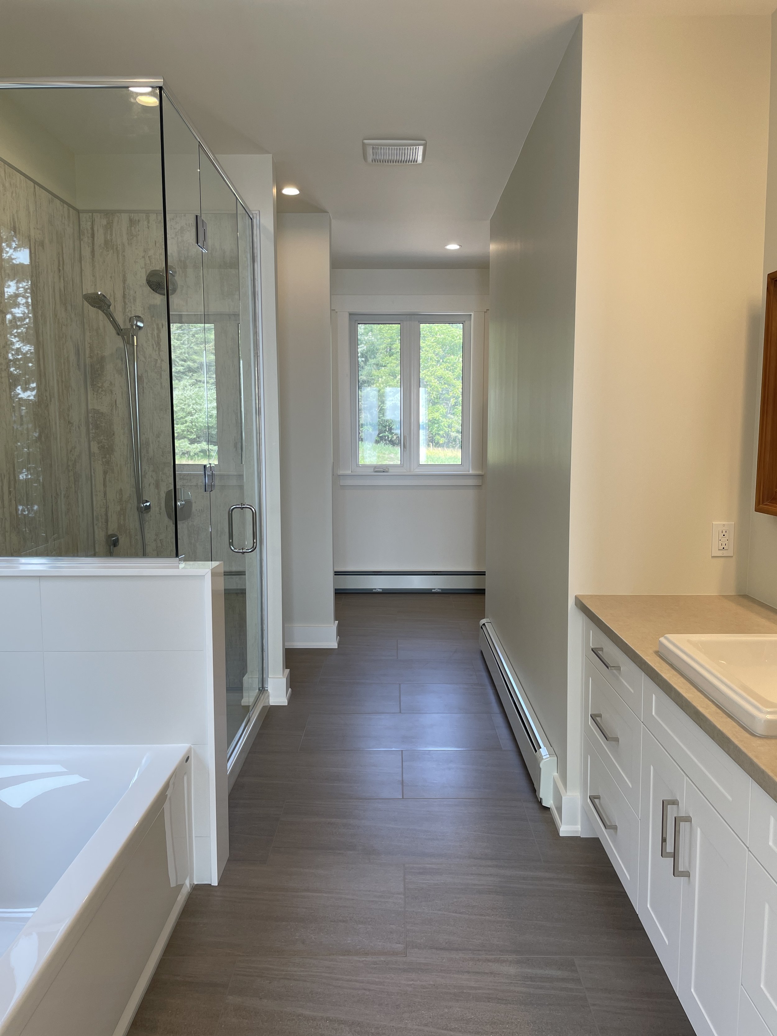 A modern bathroom with a glass shower enclosure, a large window with a view of green trees, a white bathtub, and white cabinetry with a beige countertop.
