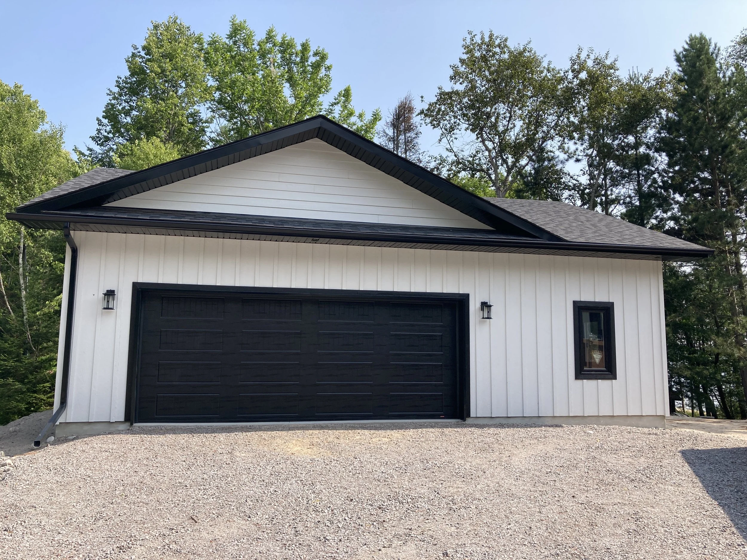 White garage with black door and black window trim, gravel driveway, surrounded by green trees and under a clear blue sky.