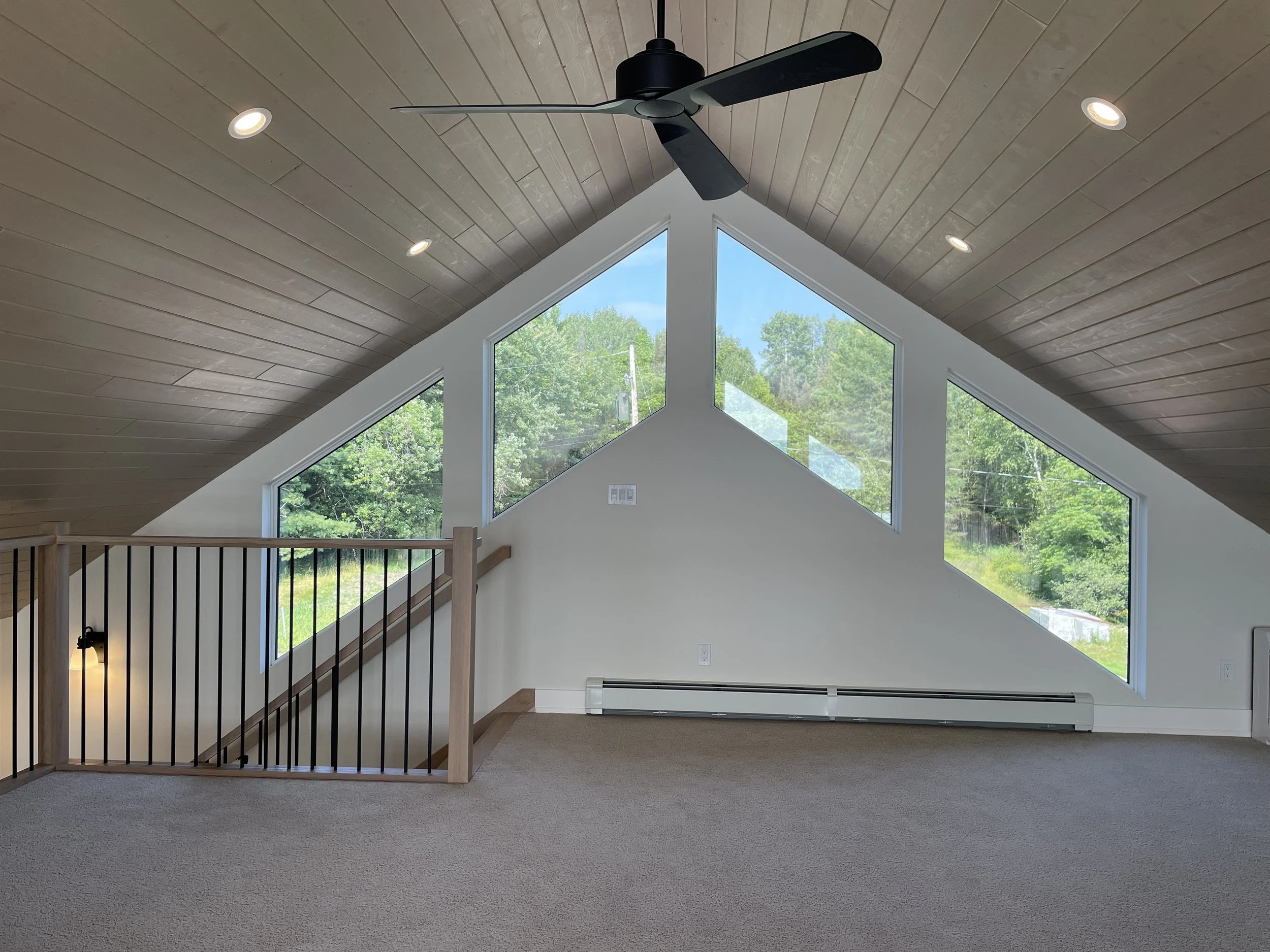Interior view of a room with a vaulted ceiling, three large triangular windows showing green trees outside, ceiling fan, recessed lighting, beige carpet, and a wooden railing leading downstairs.