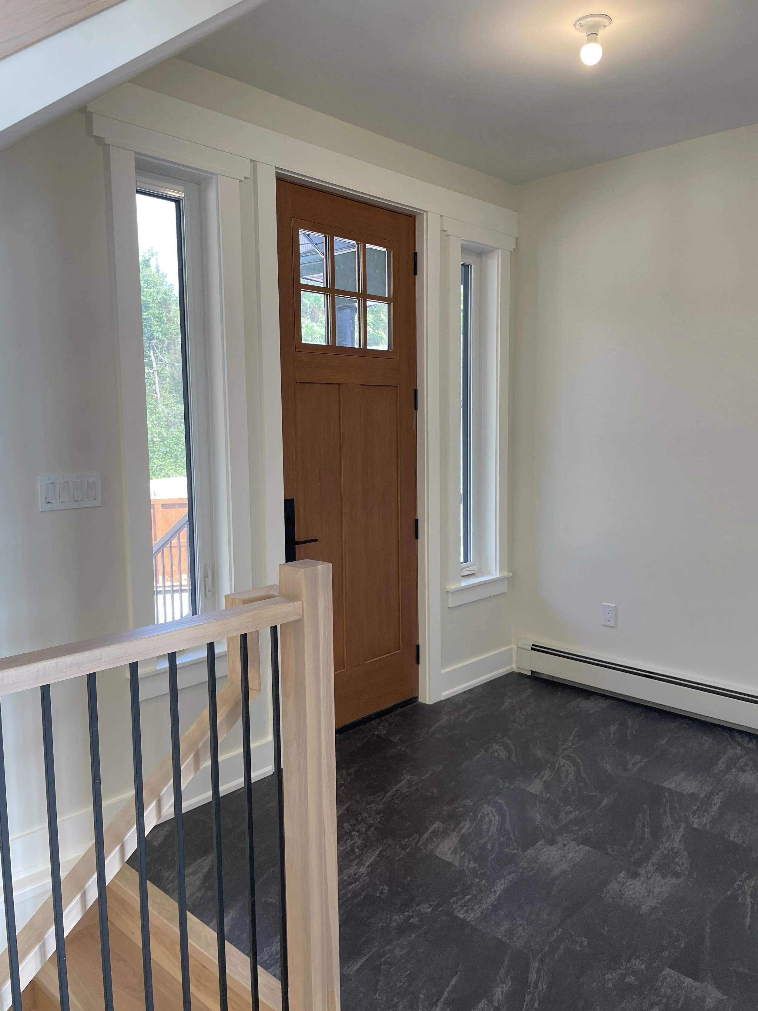 Interior view of a house entryway with a wooden door, two narrow windows on each side, a staircase with wooden handrail and black metal balusters, dark floor tiles, white walls, and ceiling light.