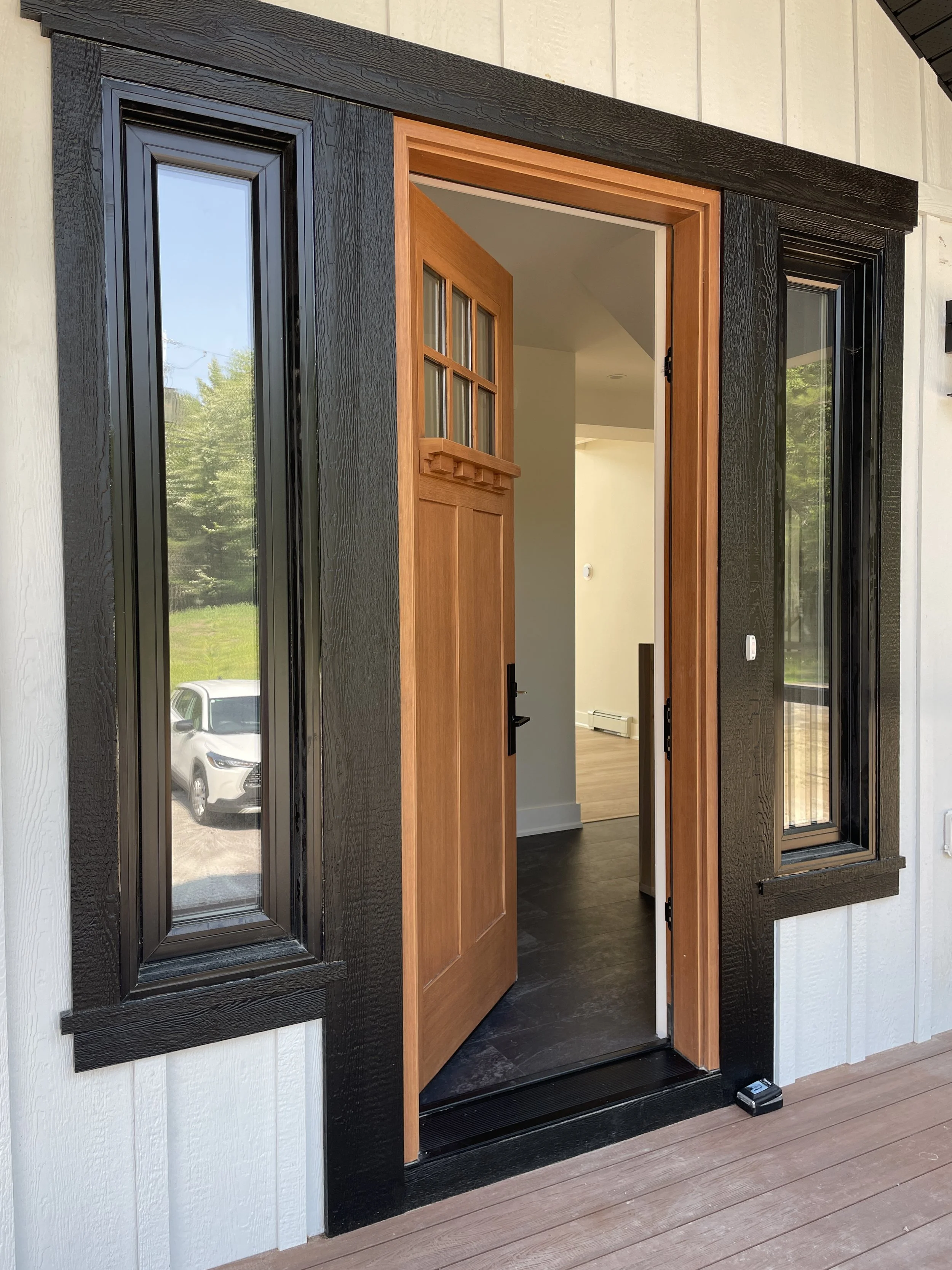 Open wooden front door with side and top windows, black framing, leading into a house with hardwood floors and white walls.