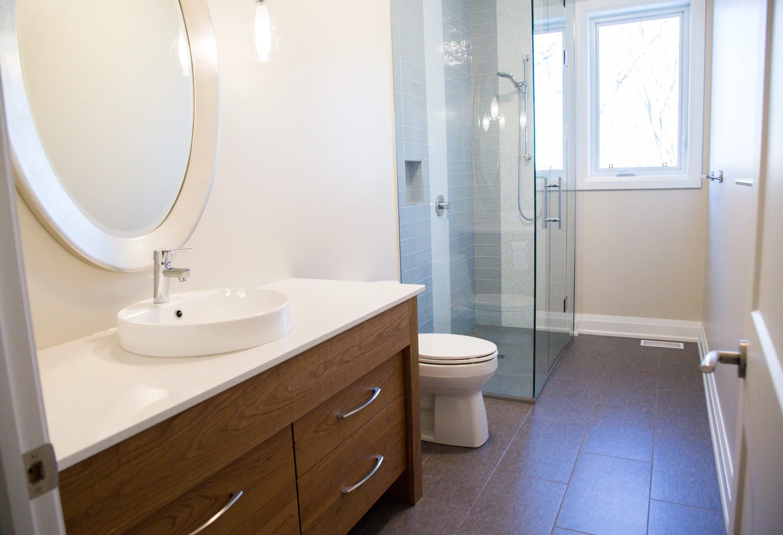 Modern bathroom with wooden vanity, oval mirror, white vessel sink, glass shower with blue tiles, toilet, and a large window with outside view.