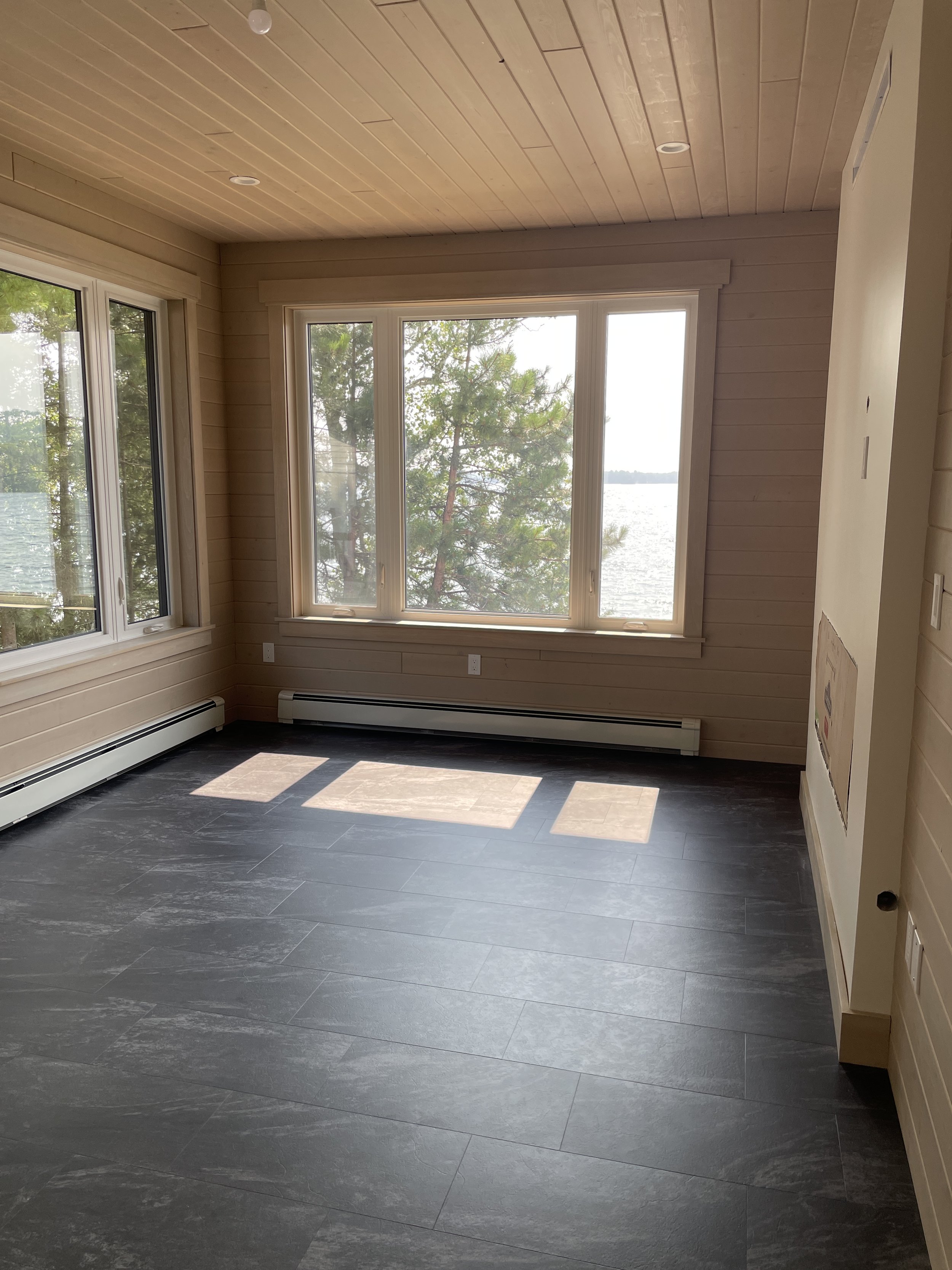 An empty room with large windows providing a view of water and trees outside. The room has wood paneling on the walls and ceiling, dark gray tiled flooring, and a baseboard heater running along the base of the interior walls.