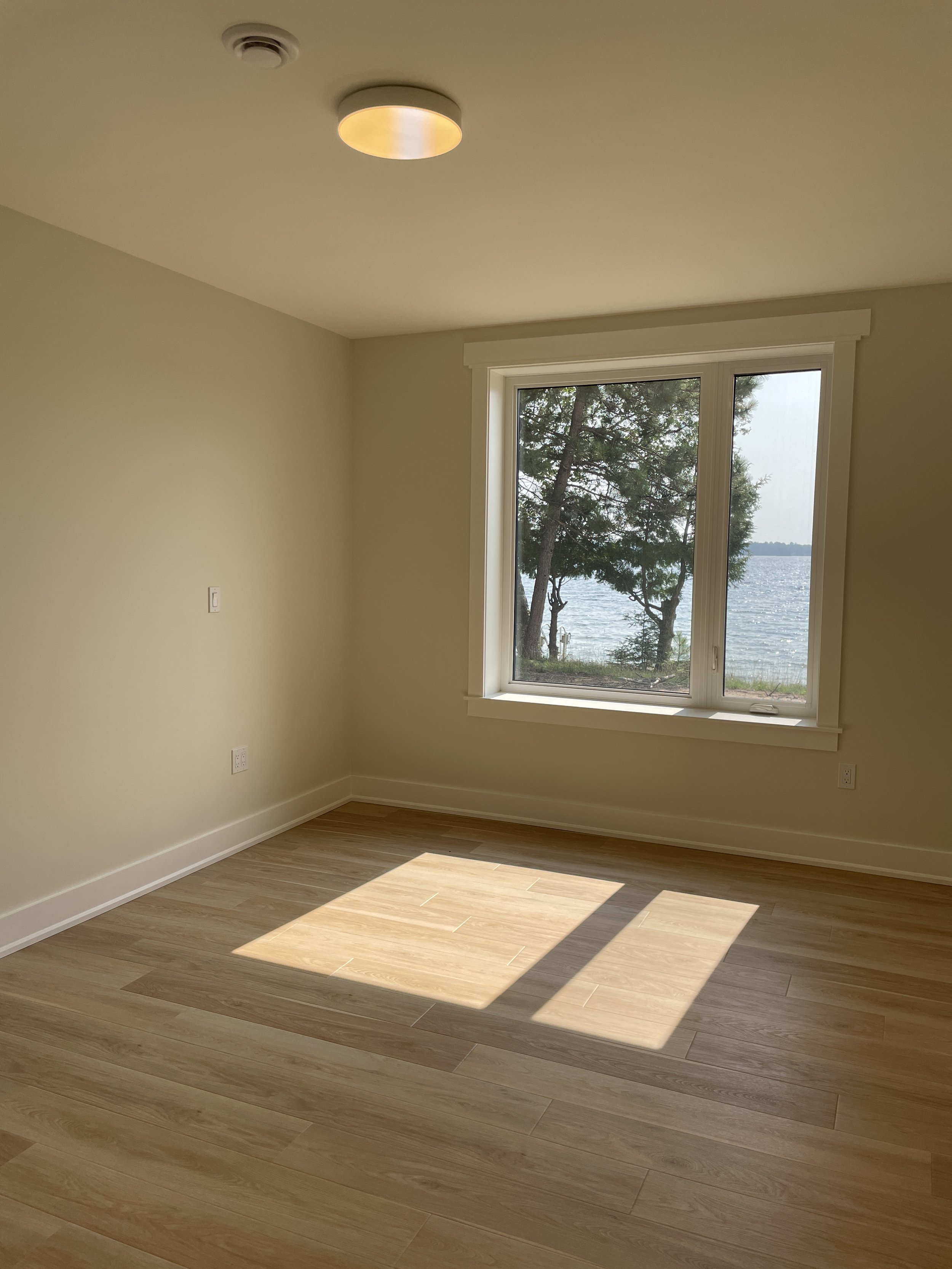 Empty room with wood flooring, beige walls, large window showing trees and water outside, ceiling light, and baseboard trim.