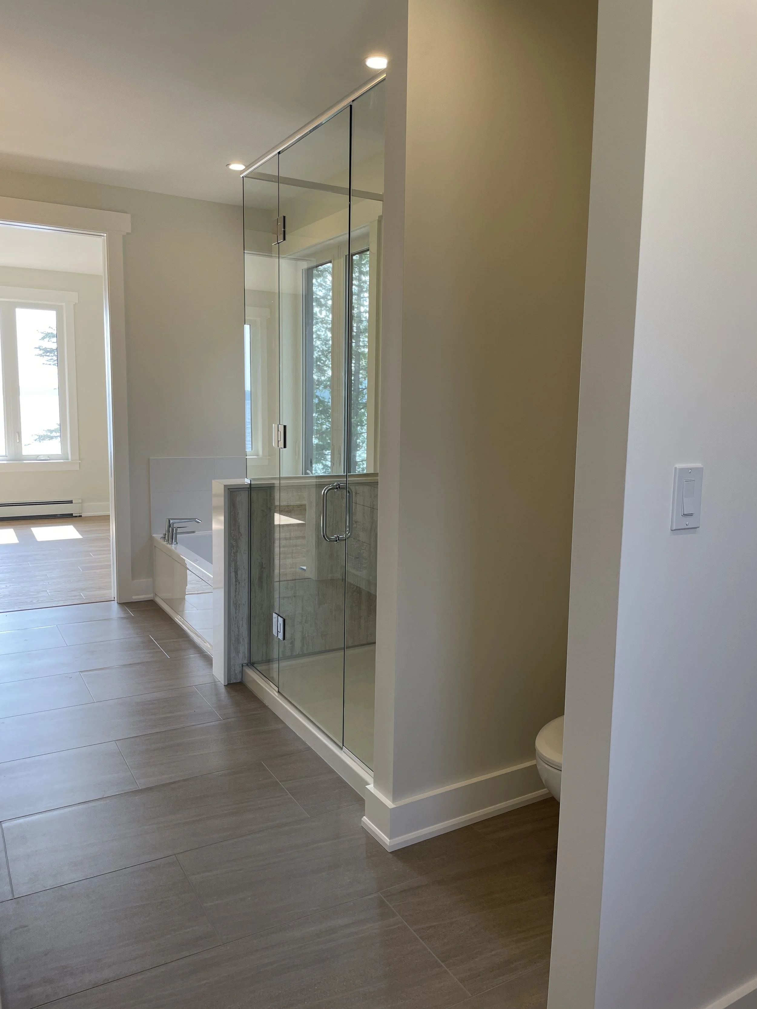 Modern bathroom with a glass shower enclosure, bathtub, and a partial view of a toilet in a small cubicle. Light-colored walls and wood-look tile flooring.