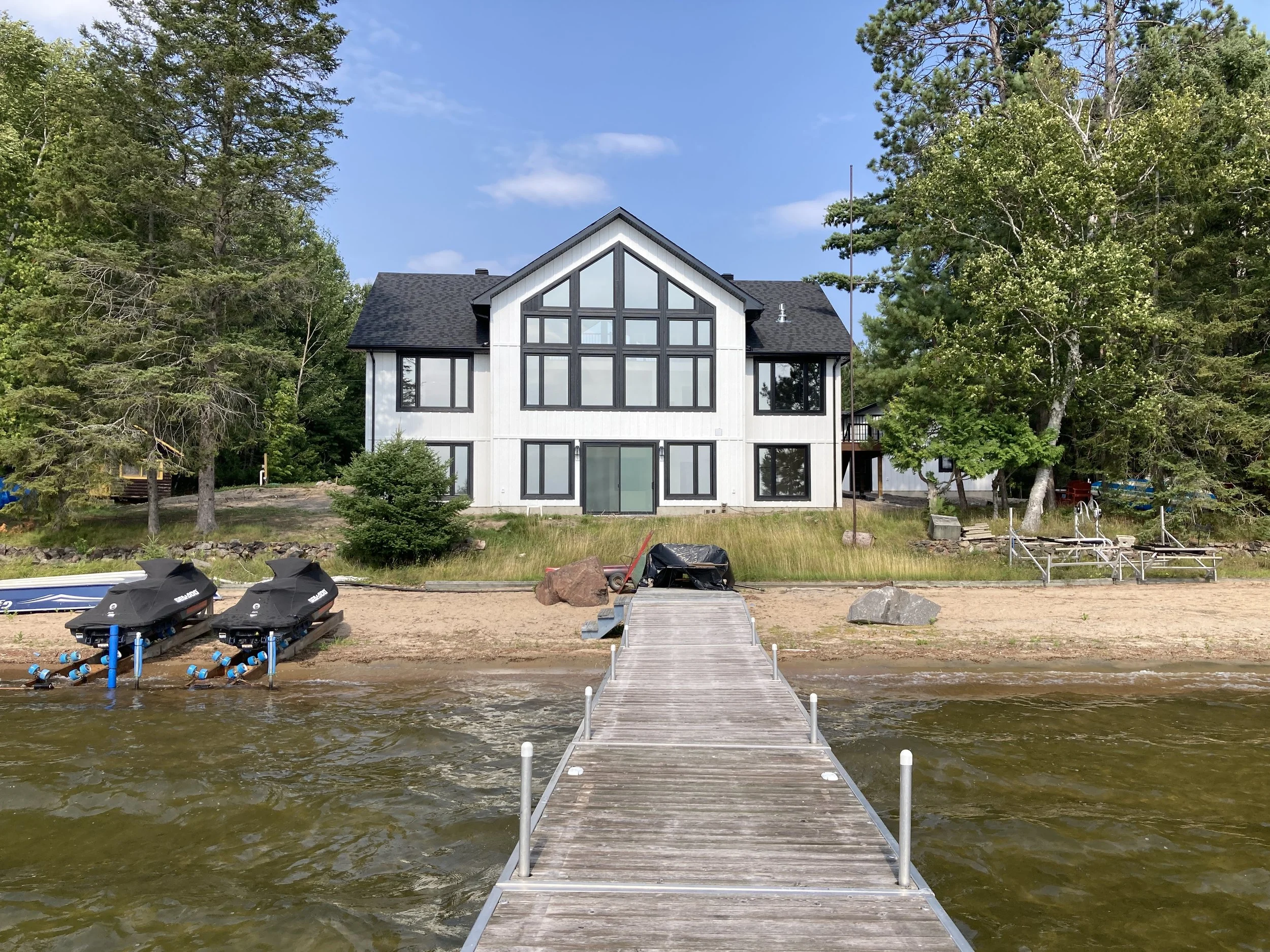 A white house with black trim facing a dock on a lake, surrounded by trees, with a sandy beach and boats in the water.