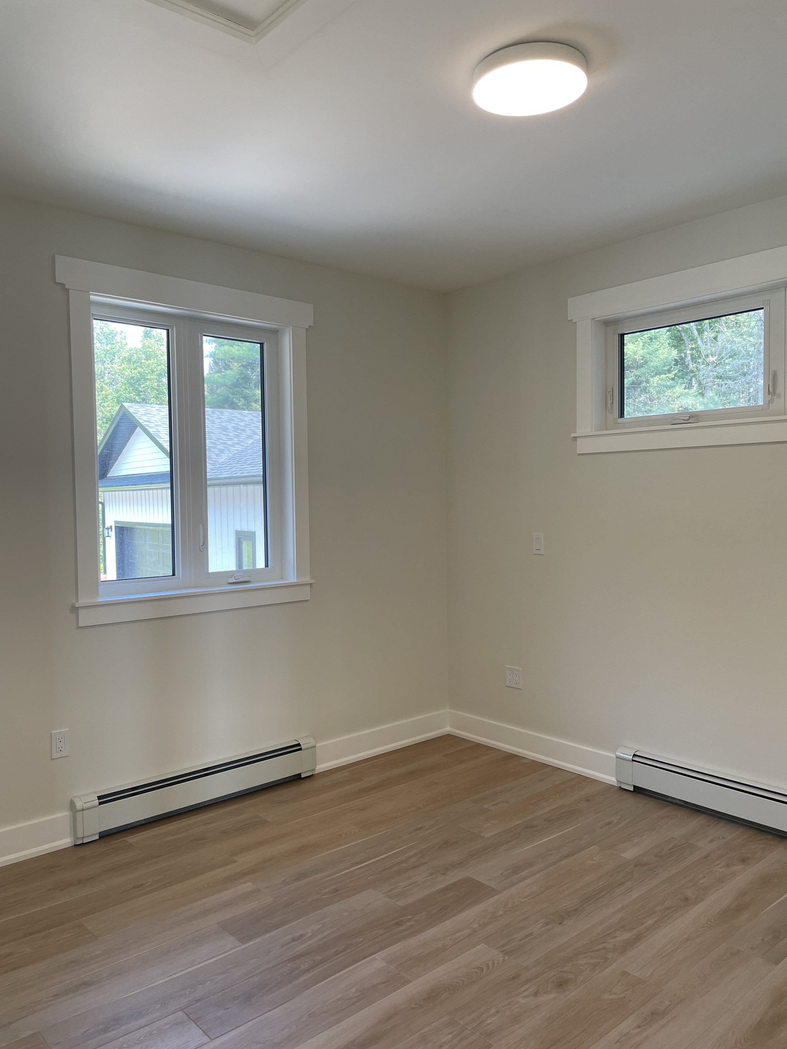 Empty room with two windows, hardwood floors, white walls, baseboard heating, and a ceiling light
