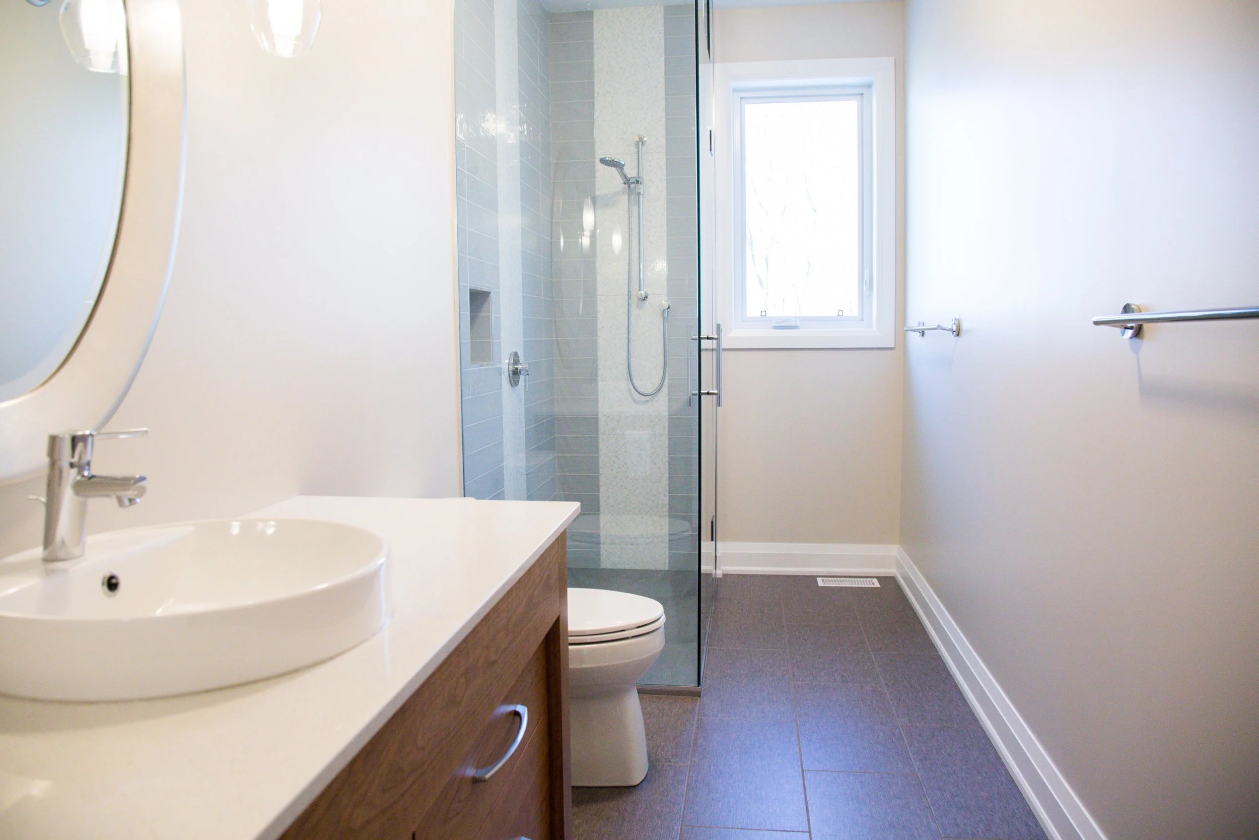 A modern bathroom with a vanity, a toilet, and a walk-in shower with a glass door and a window.