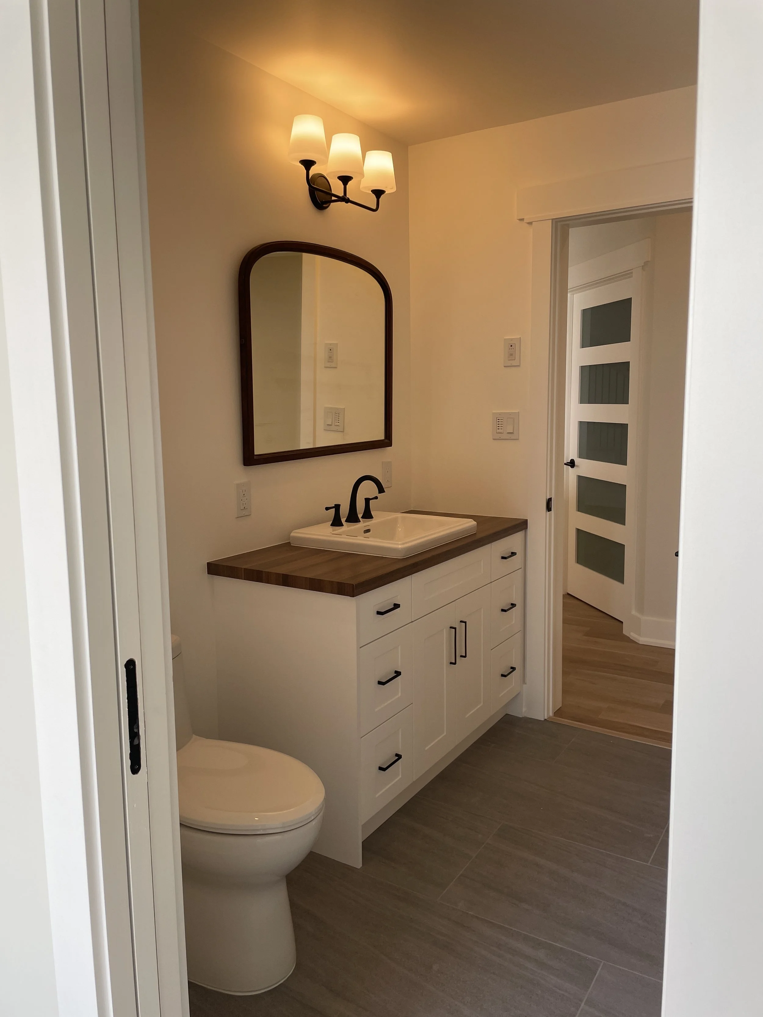 A modern bathroom with a white vanity, a rectangular sink, and a black faucet. There is a large mirror above the sink, a wall-mounted light fixture with three lamps, and a door with frosted glass panels. The bathroom has wood-look flooring.