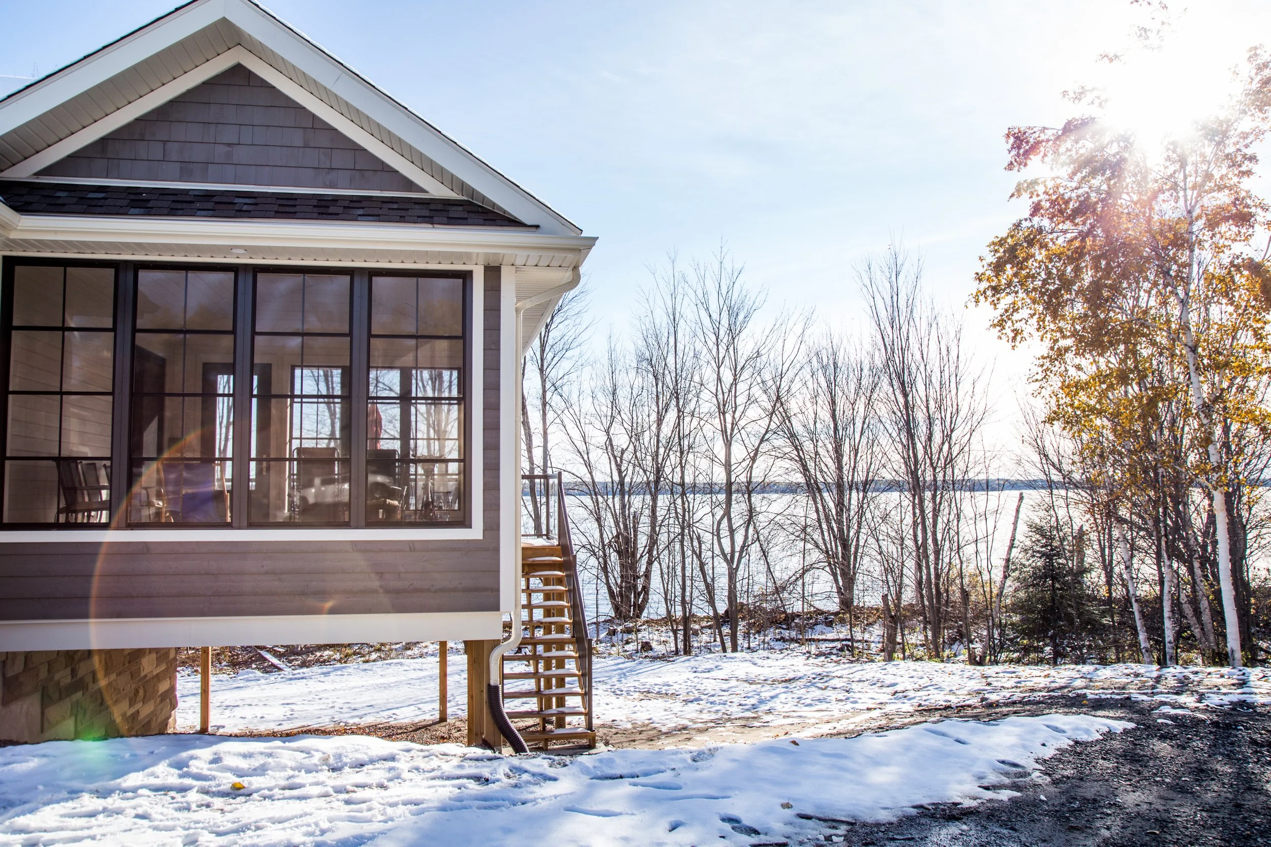 A house with a porch overlooking a snowy landscape with leafless trees, a lake, and a bright sun in the sky.