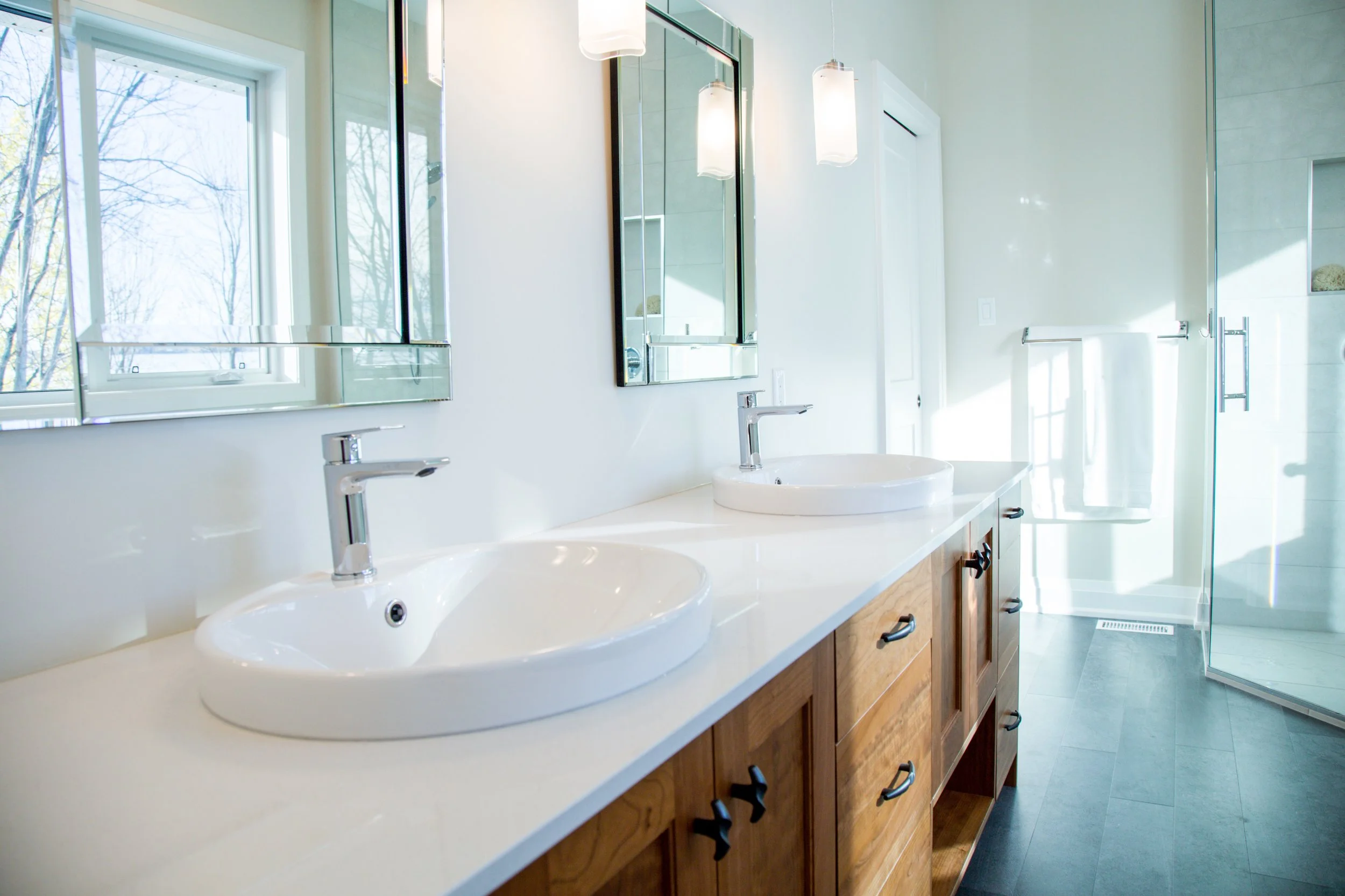 Modern bathroom featuring double sinks, large mirrors, wooden cabinet, hanging lights, a towel rack with towel, and a glass-enclosed shower with a window showing tree branches outside.