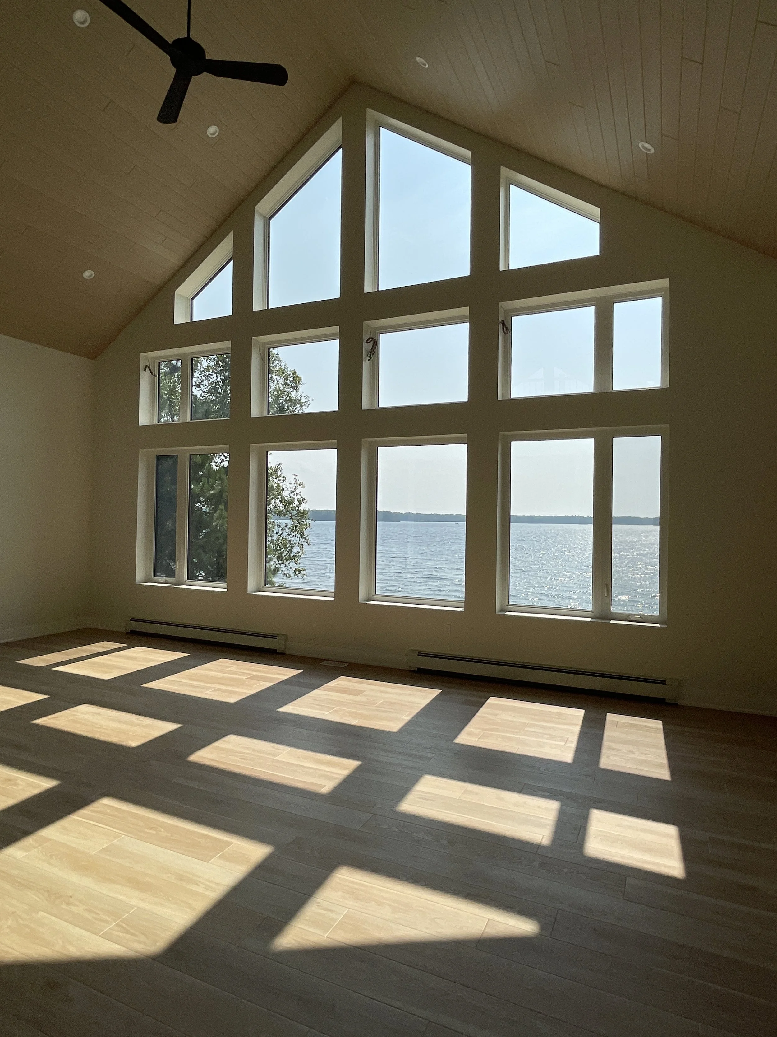 Empty living room with large multi-pane windows overlooking a lake, sunlight casting shadow patterns on the wooden floor, vaulted wooden ceiling with ceiling fan.