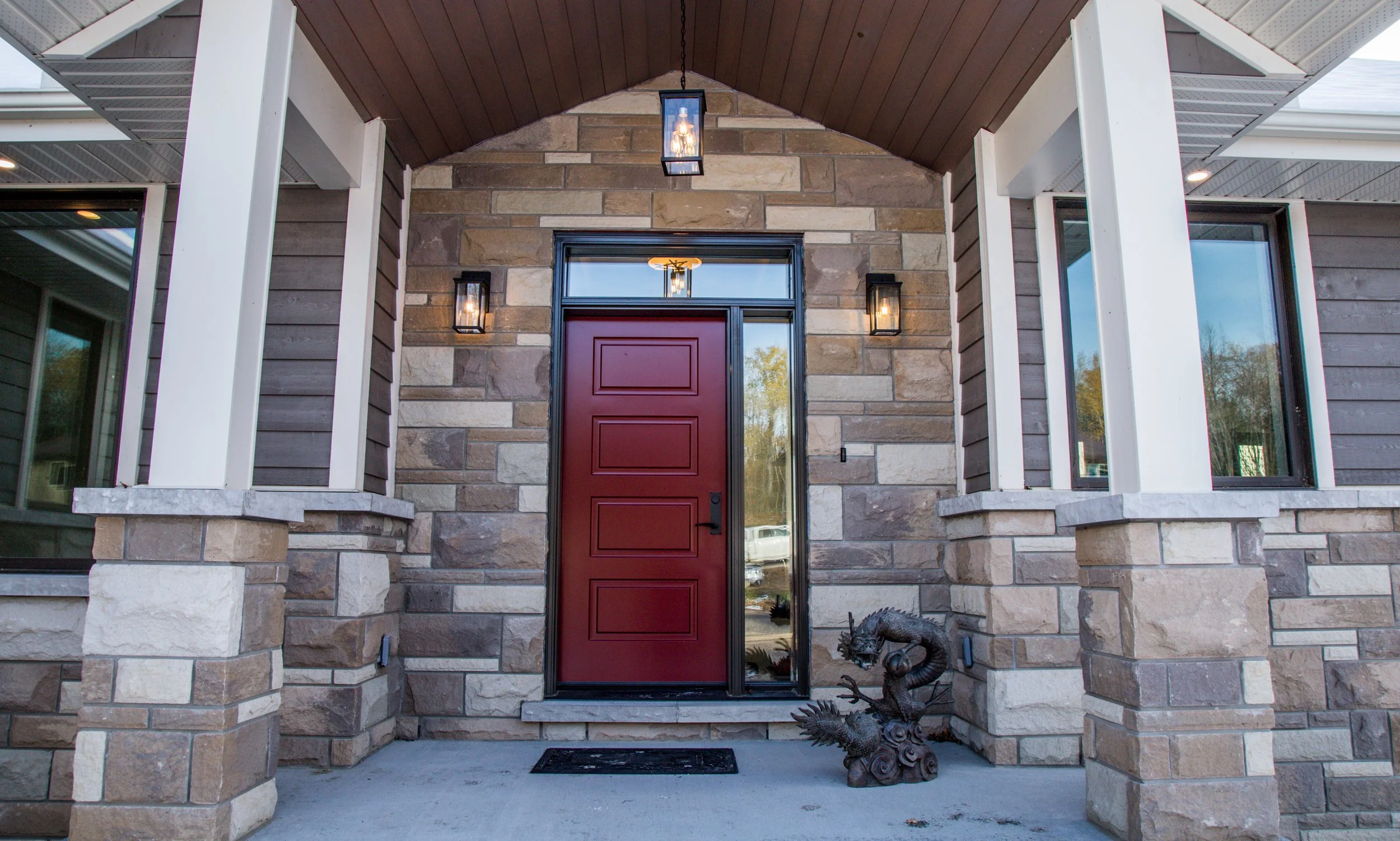 Front door of a house with a red door, stone and wood exterior, two lantern-style wall lights, a hanging lantern ceiling light, and a decorative dragon sculpture near the door.