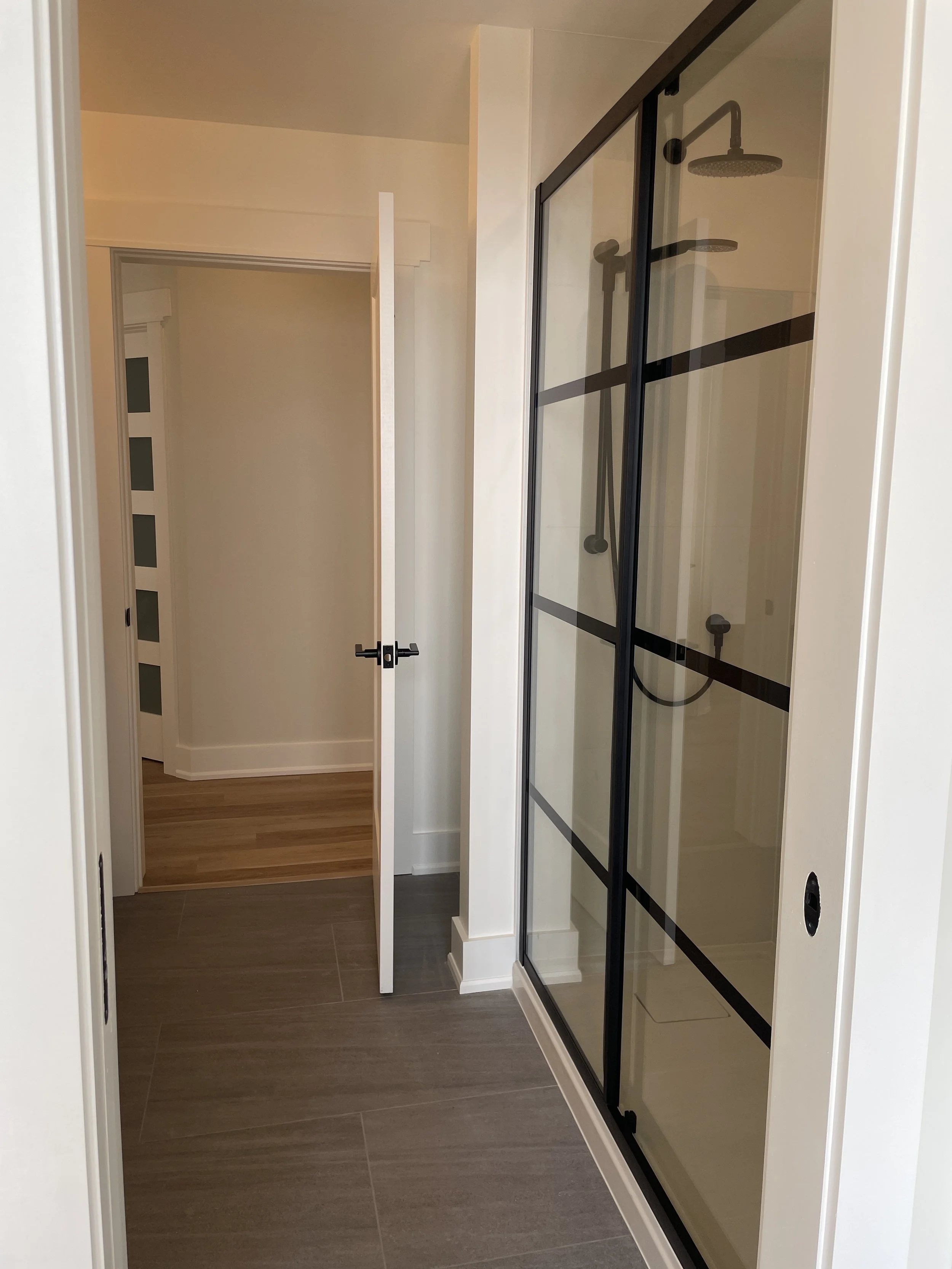 Bathroom with a glass shower enclosure, a closed white door, beige walls, and wooden flooring.