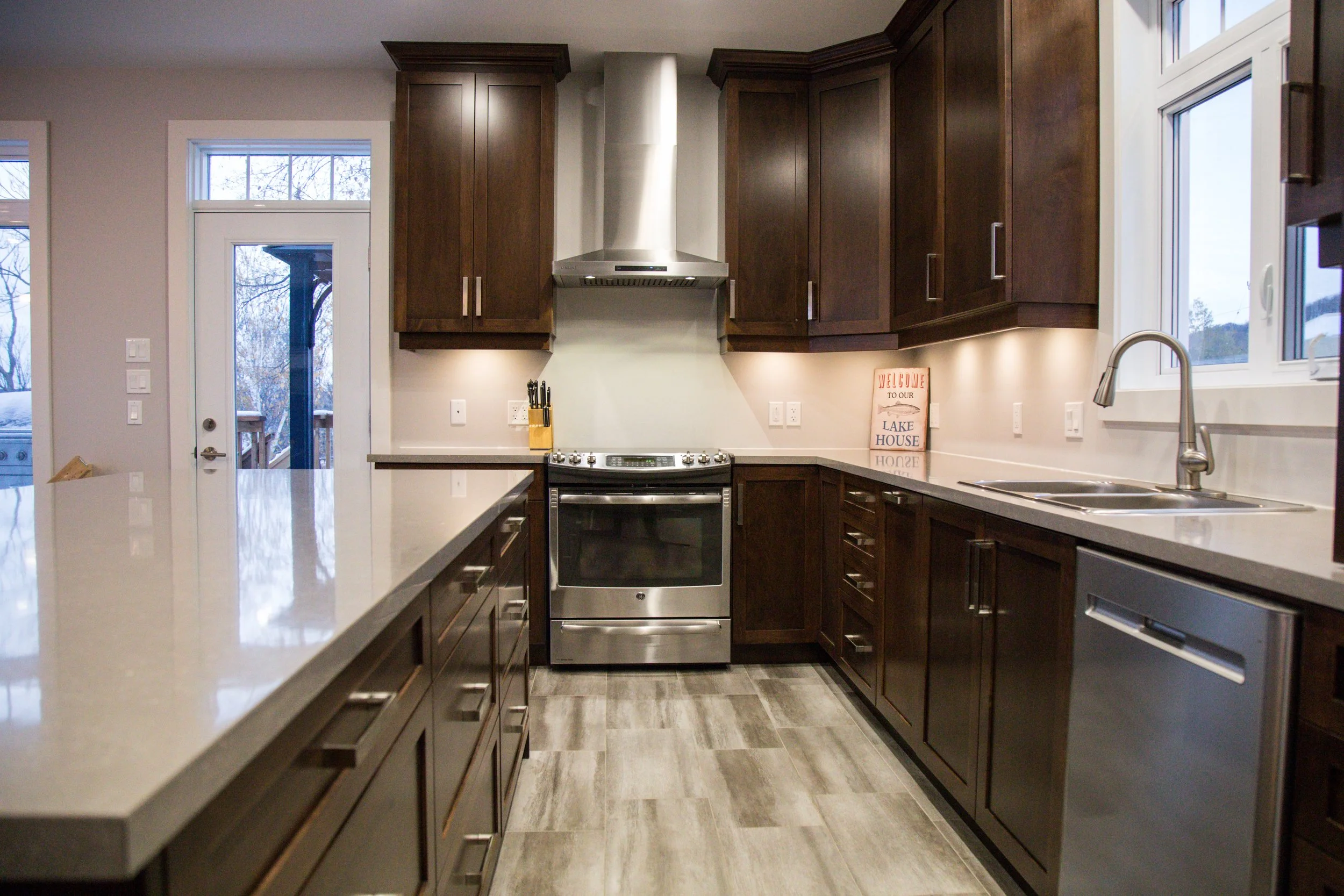 Modern kitchen with dark wooden cabinets, stainless steel stove, dishwasher, and silver range hood. Light countertops, a double sink, and a window overlooking the outside. A sign on the counter reads 'Welcome to our Lake House.'