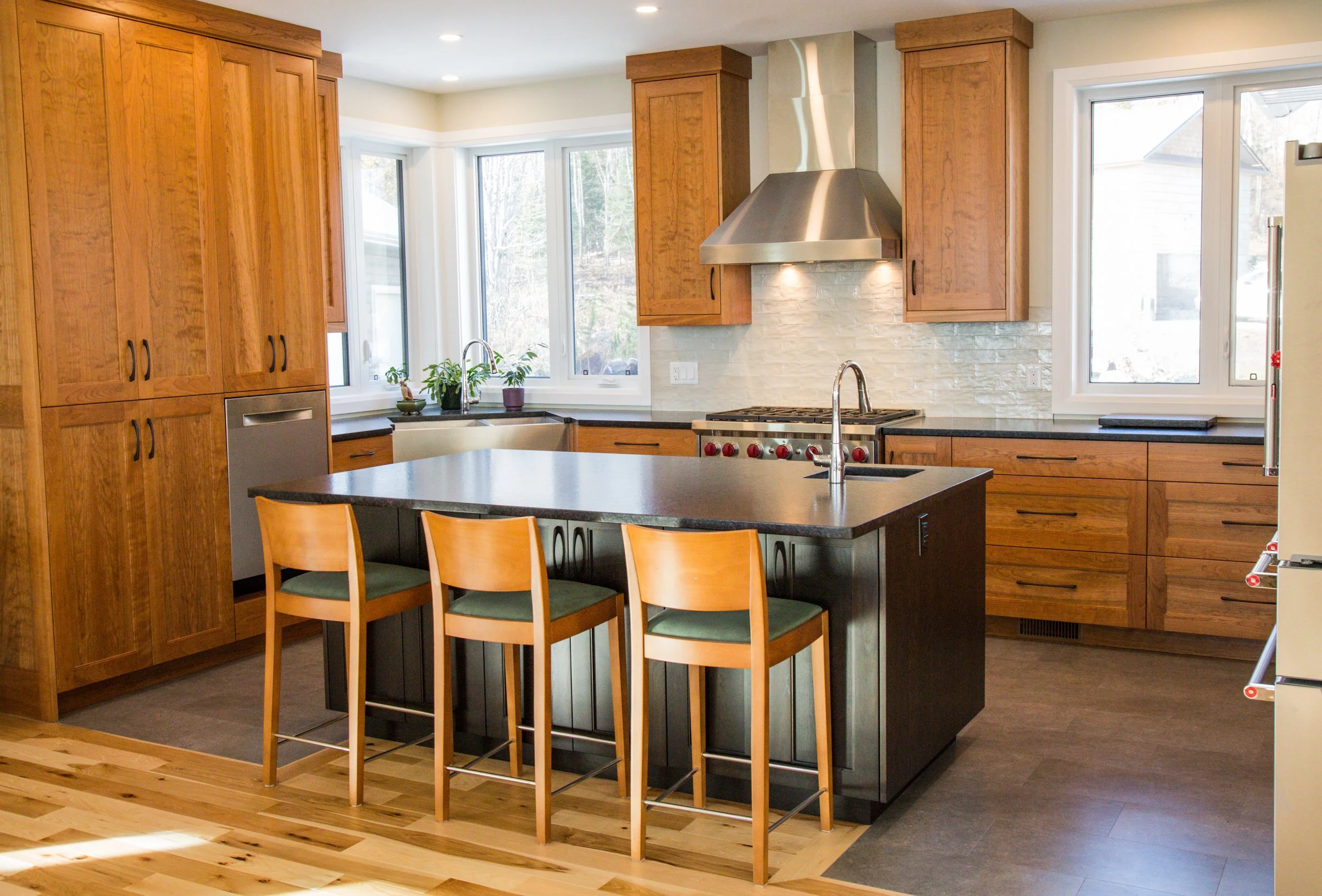 Modern kitchen with wooden cabinets, a black island with three chairs, stainless steel appliances, white brick backsplash, windows, and a hardwood floor.