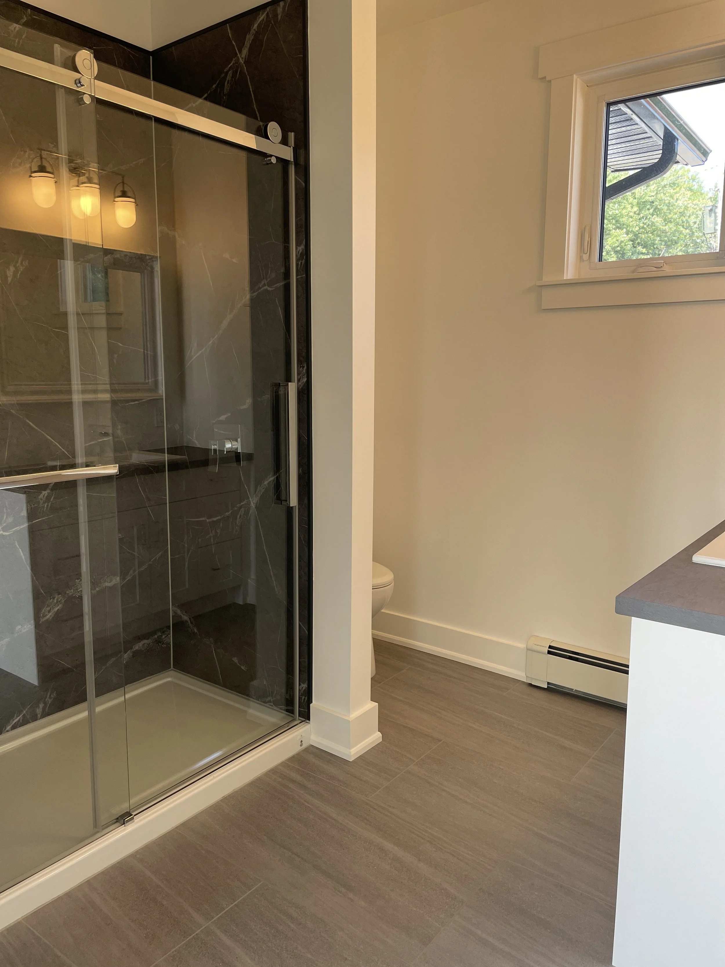 A bathroom with a glass shower enclosure on the left, a small window on the right, and a toilet partially visible behind a wall. The bathroom has grayish wood-look flooring and a countertop on the right side.