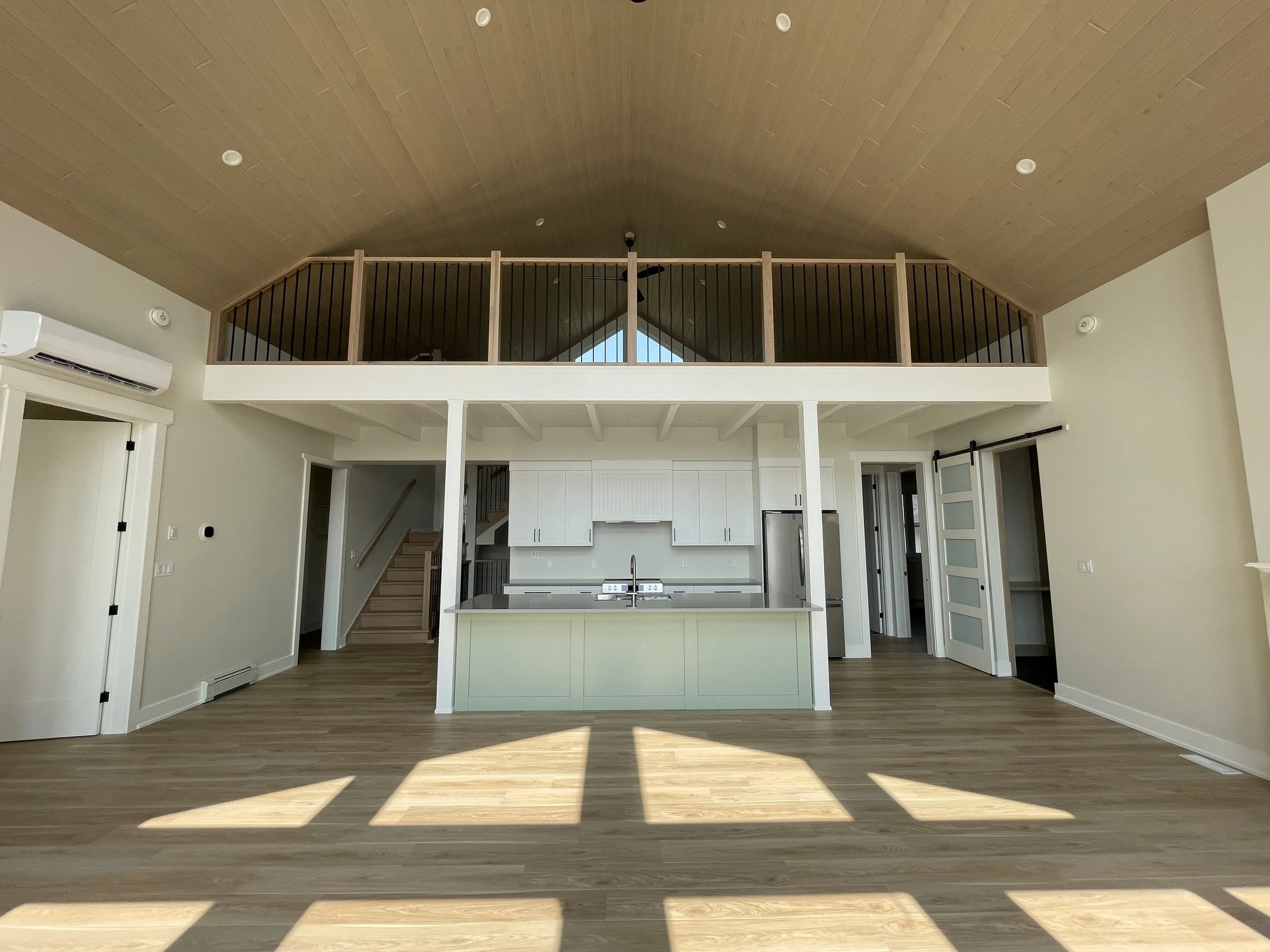Living area with a kitchen featuring a central island, white cabinets, and stainless steel appliances, including a refrigerator. Upstairs has a loft with a wooden railing and stained wood ceiling, with large windows illuminating the room.