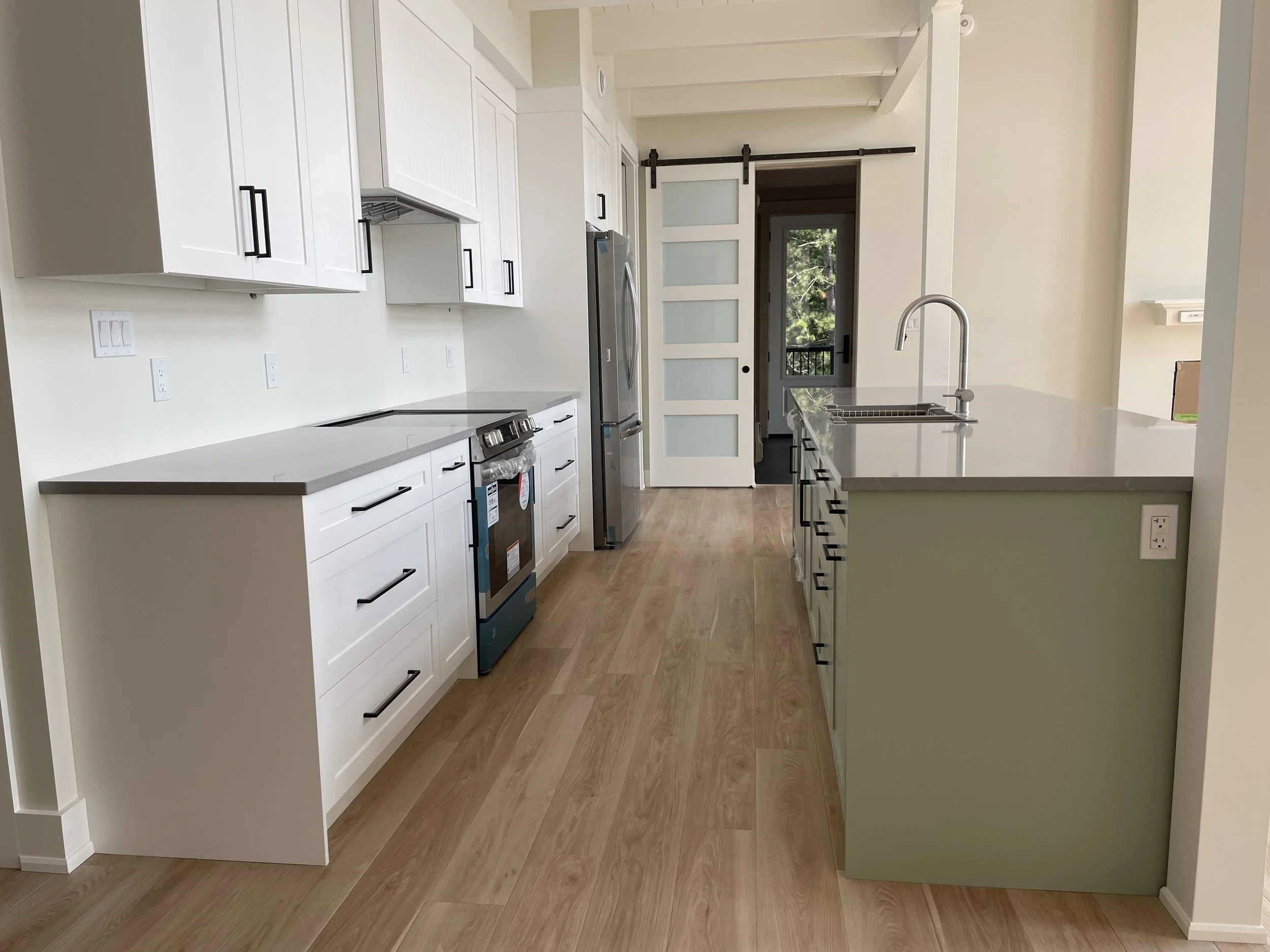 A modern kitchen with white cabinets, stainless steel appliances, and light wood flooring. There is a sliding barn door in the background leading outside or to another room, and a large kitchen island with a sink.