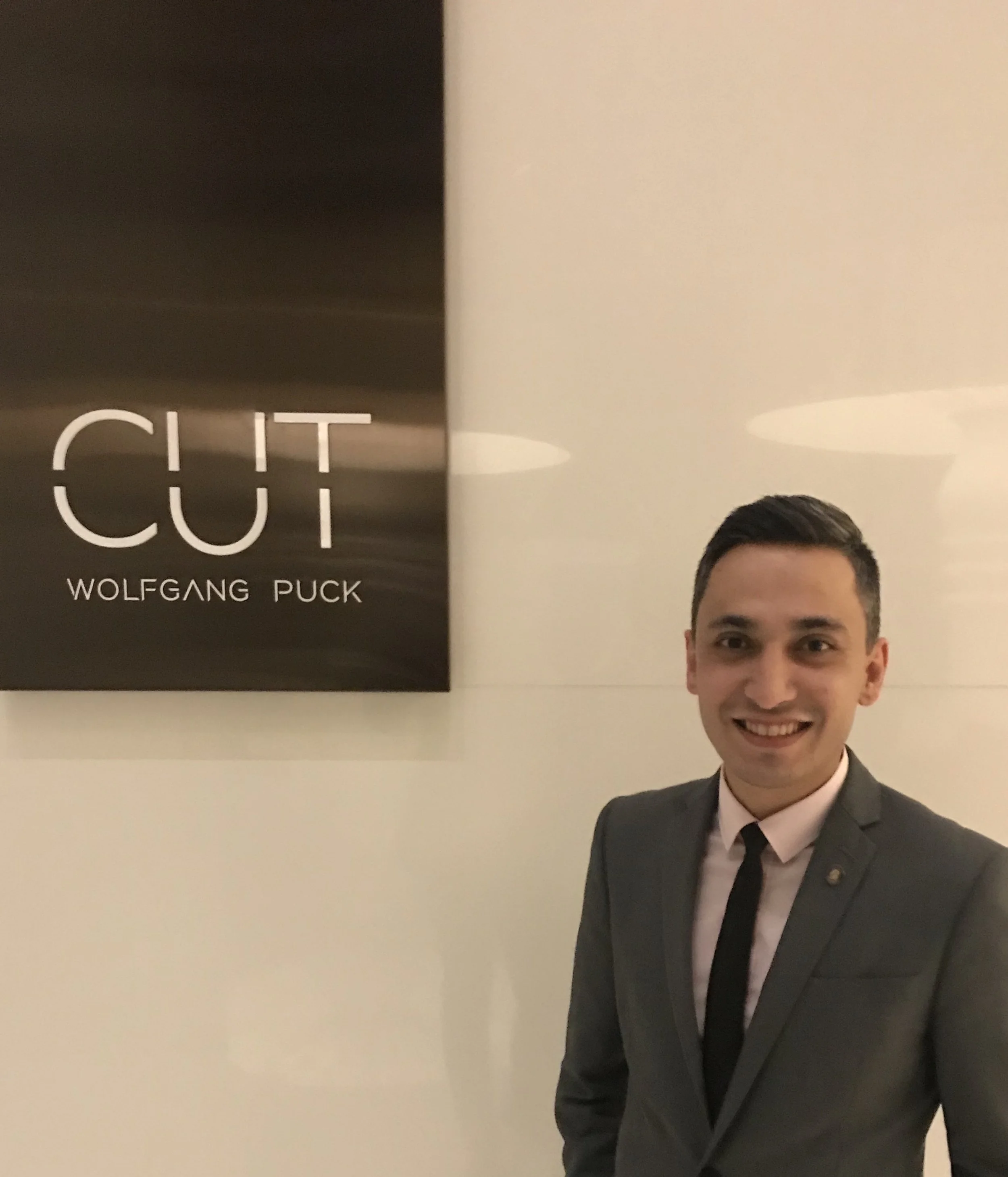 A man in a suit standing next to a poster for a play titled 'CIT' by Wolfgang Puck, smiling at the camera in an indoor setting.