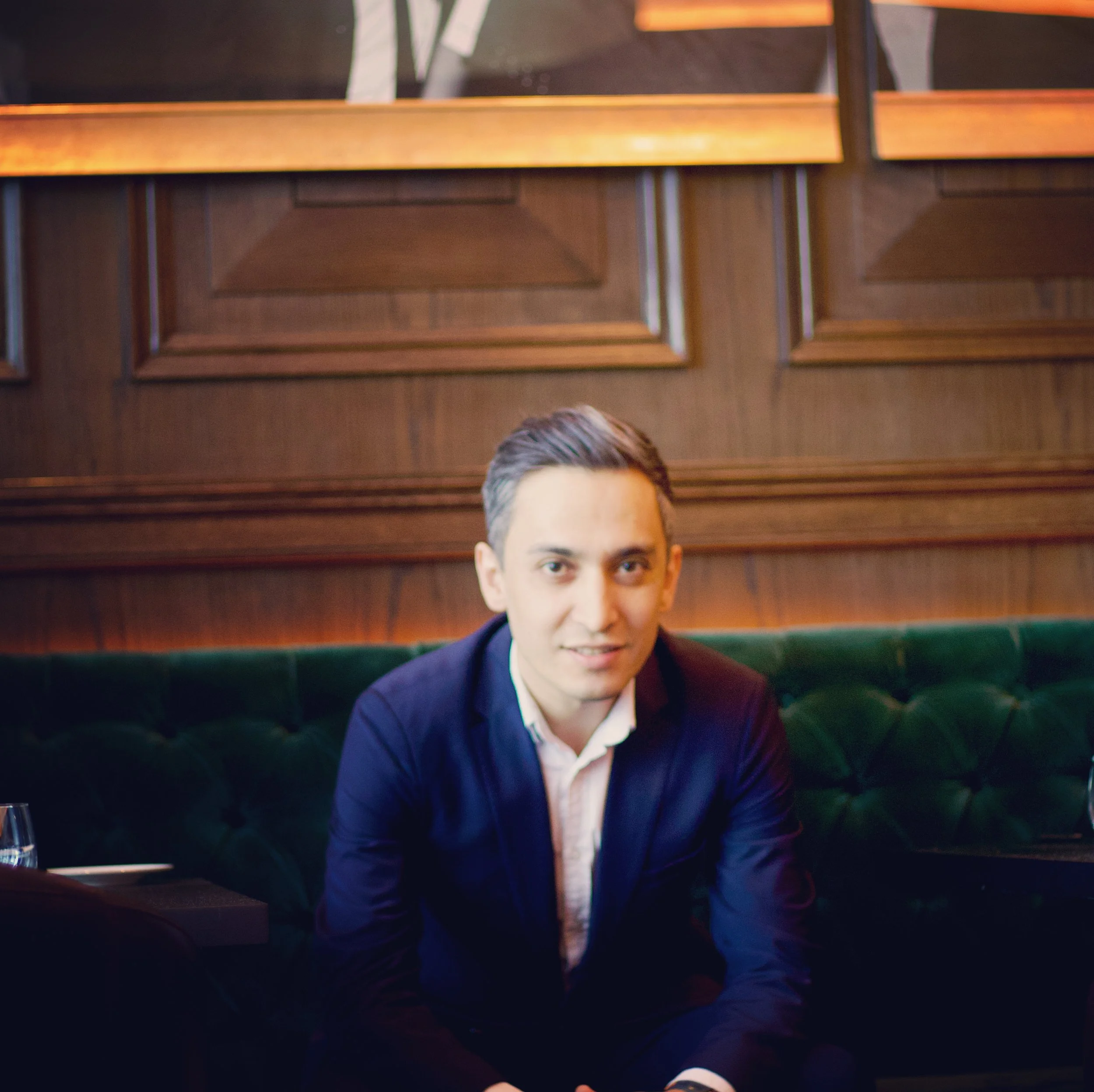 A young man with short dark hair, wearing a navy blazer and white shirt, sitting on a green velvet booth in a restaurant with wooden paneling on the walls.