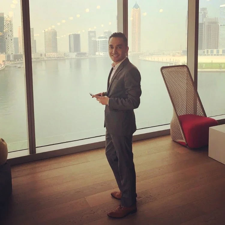 A man in a gray suit and white shirt standing indoors near large glass windows with a city skyline and water view behind him, holding a smartphone and smiling.