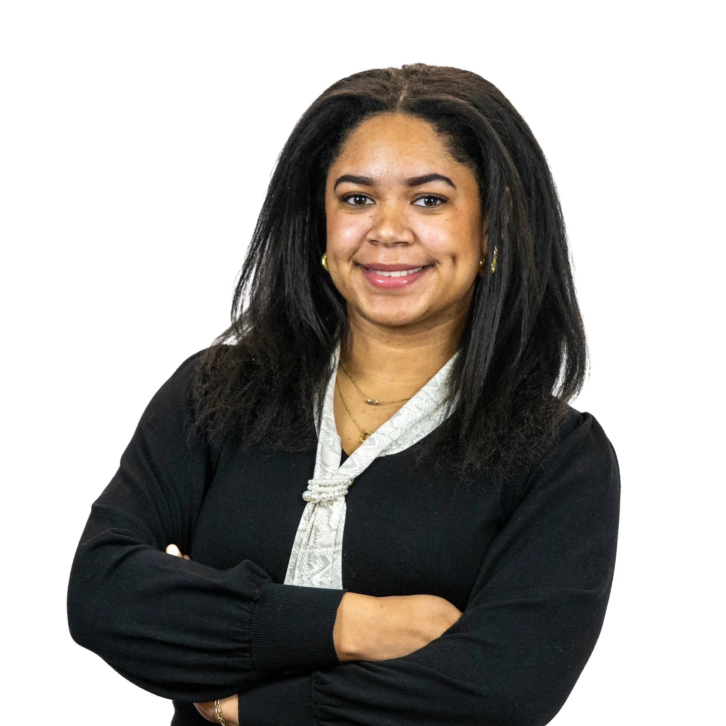 A woman with medium-length curly black hair, wearing a black blazer, white blouse with a textured pattern, and gold jewelry, standing with arms crossed against a white background.