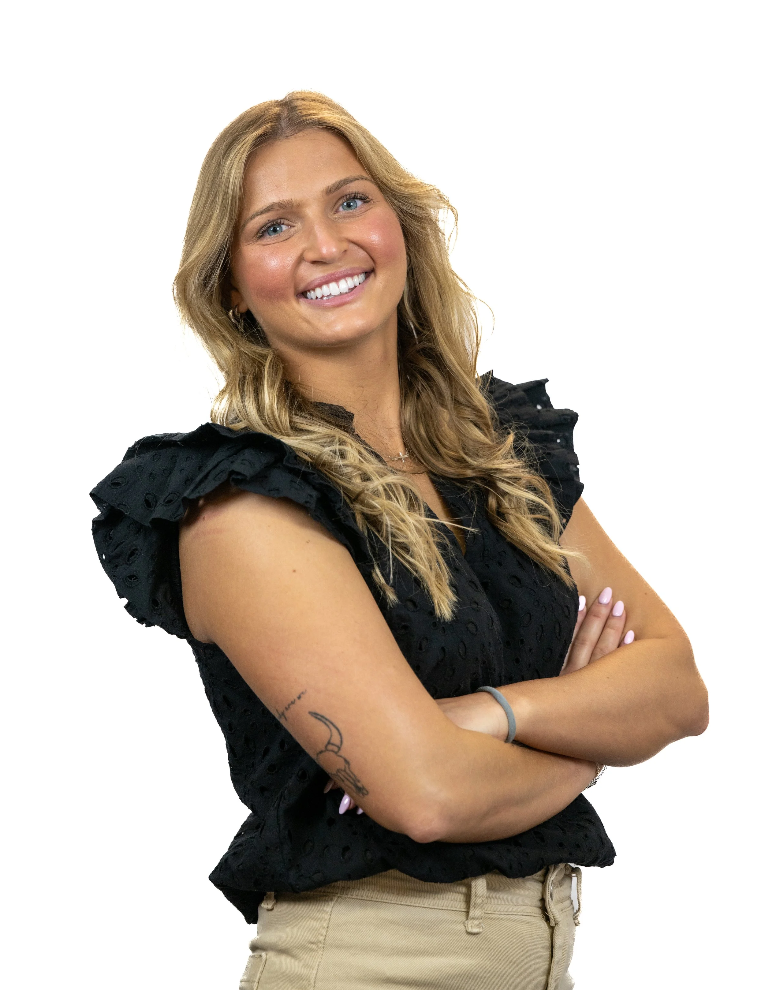 A smiling woman with long blond hair, wearing a black eyelet ruffled top, beige pants, and a tattoo on her left arm, standing with arms crossed against a plain white background.