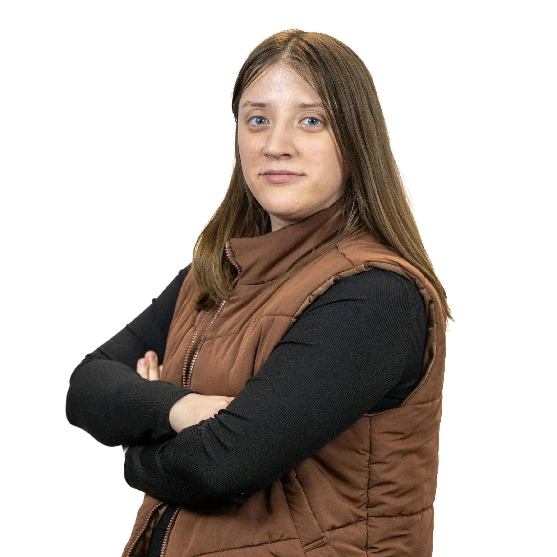 A woman with long brown hair, blue eyes, and a neutral expression, standing with arms crossed, wearing a black long-sleeve top and a brown quilted vest, against a white background.