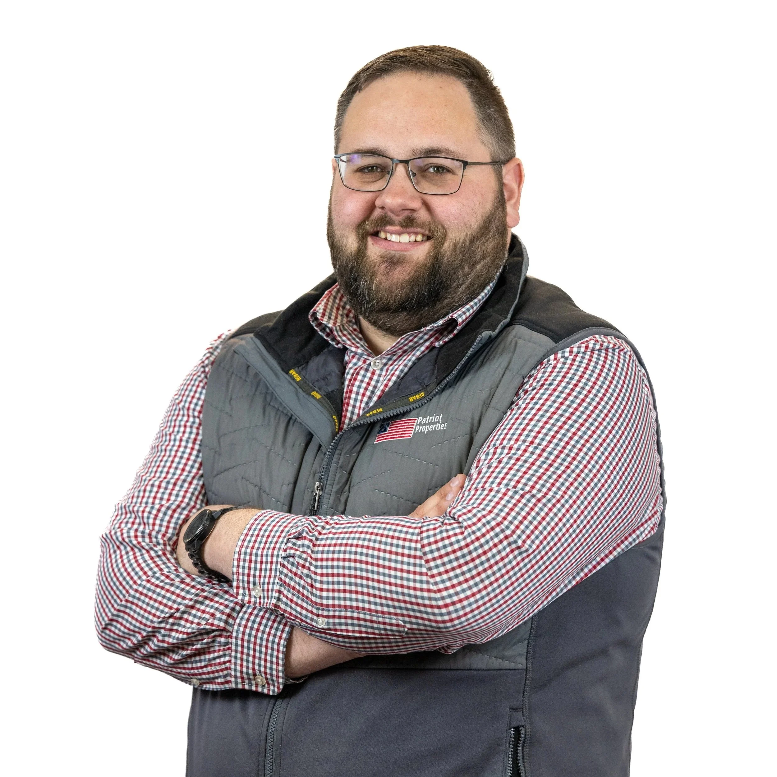Man with glasses and beard smiling, wearing a checkered shirt and gray vest, standing with arms crossed against a plain white background.