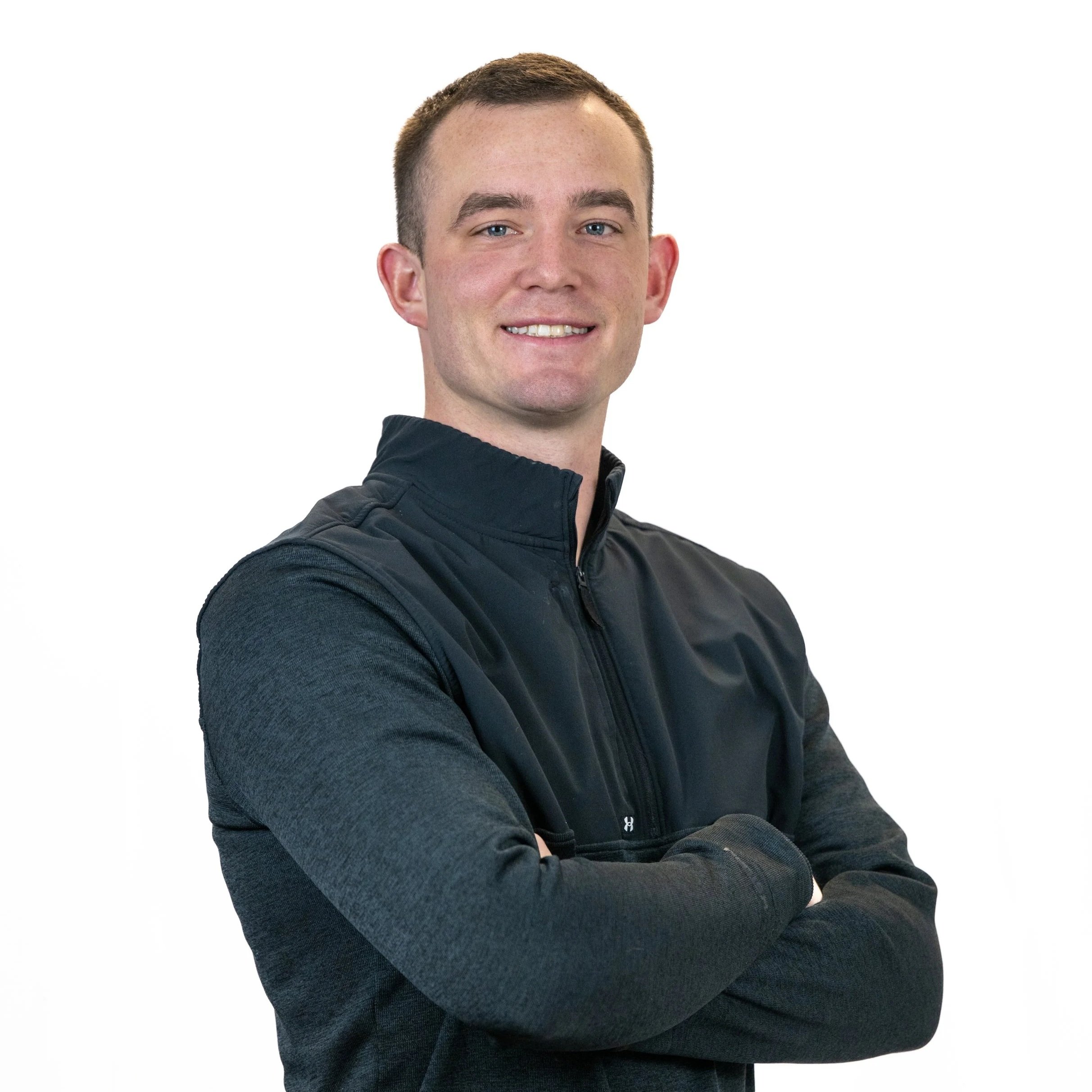 Young man with short brown hair, smiling and looking at the camera, wearing a black athletic zip-up jacket with his arms crossed, against a plain white background.