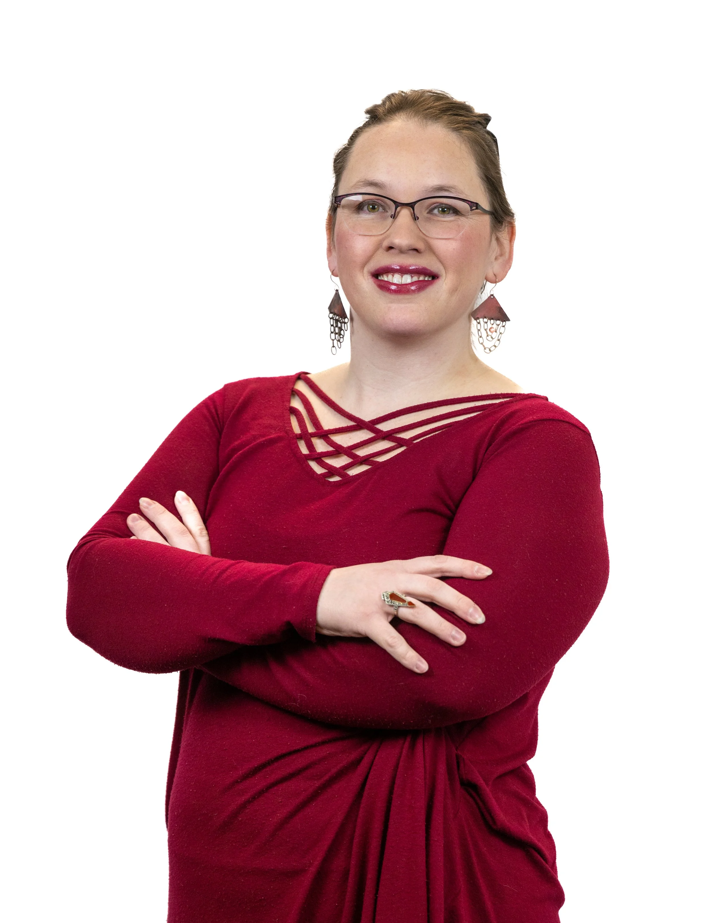 A woman with glasses and earrings wearing a red dress with cross strap neckline, standing with arms crossed and smiling