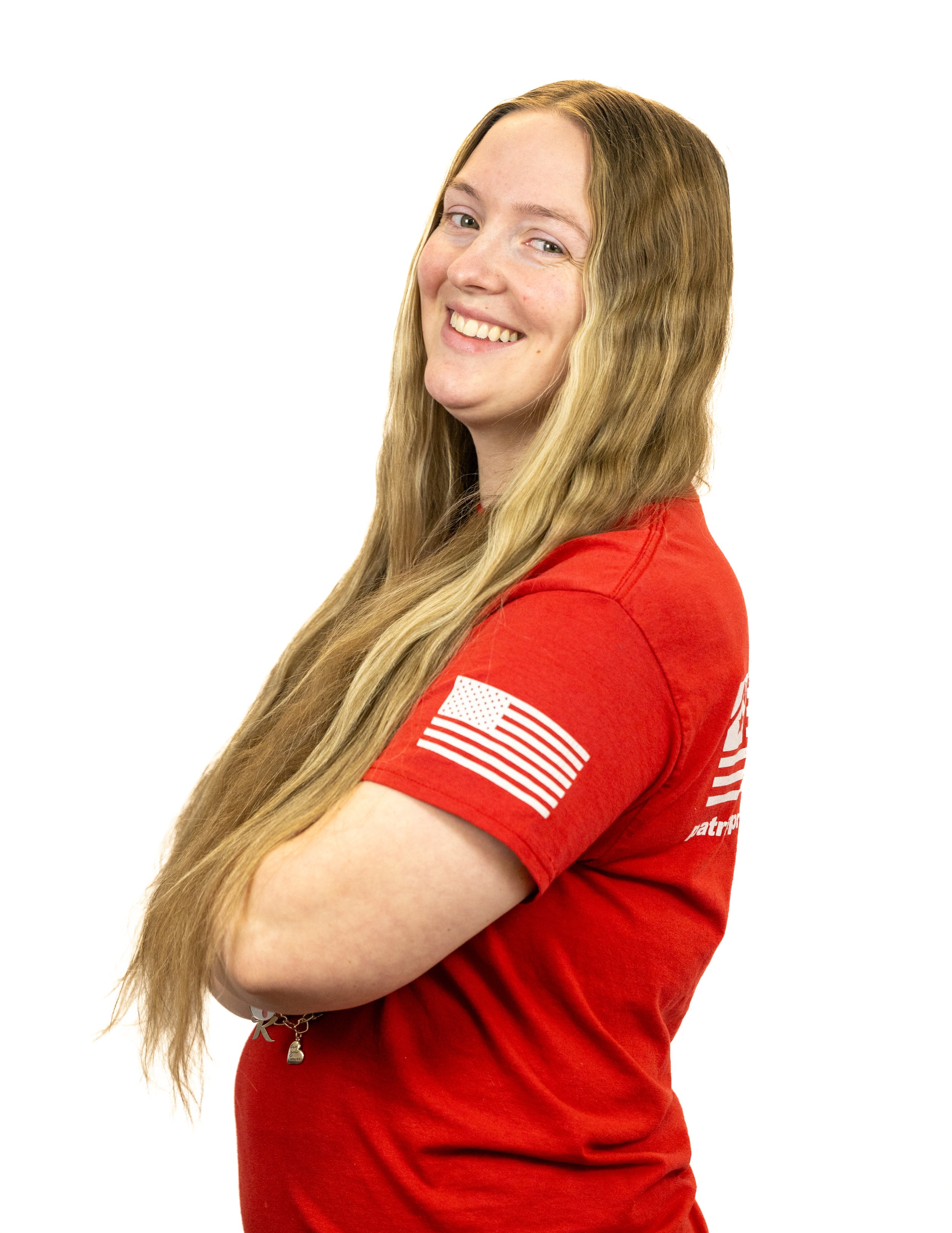 A young woman with long blonde hair smiling and wearing a red T-shirt with an American flag patch on the sleeve, standing with arms crossed against a white background.