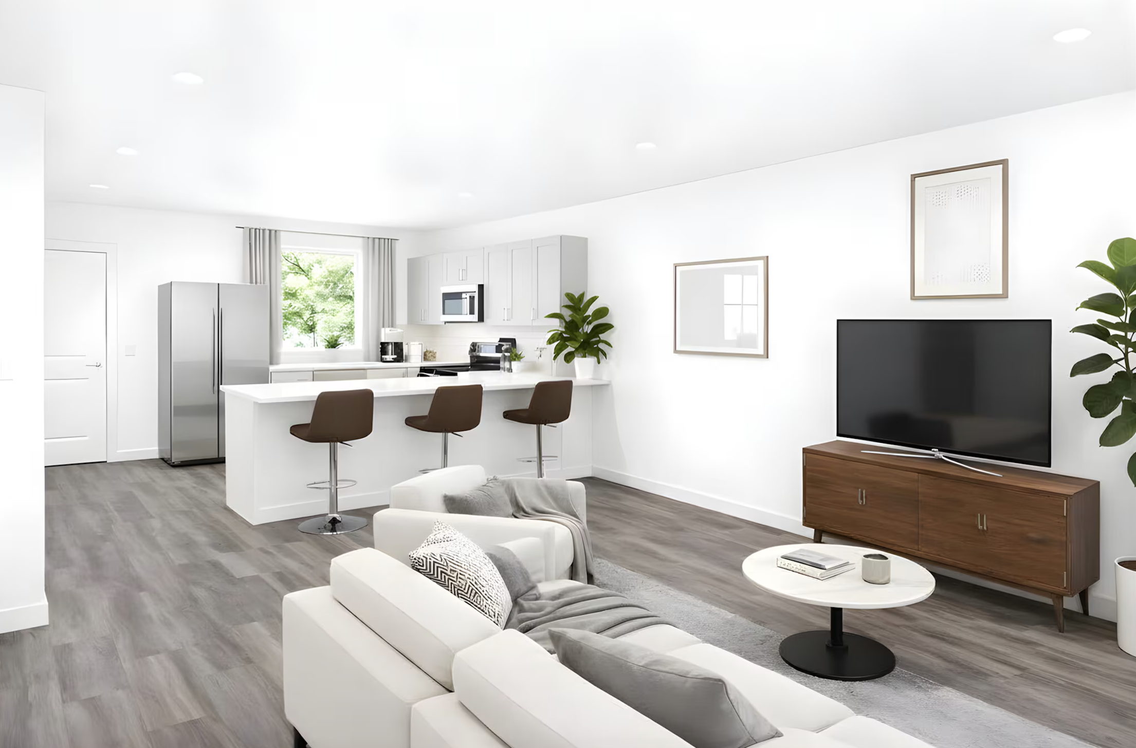 Modern open-concept living room and kitchen with white walls, grey wood flooring, white and brown furniture, a large flat-screen TV on a wooden TV stand, green plants, and framed artwork on the wall.
