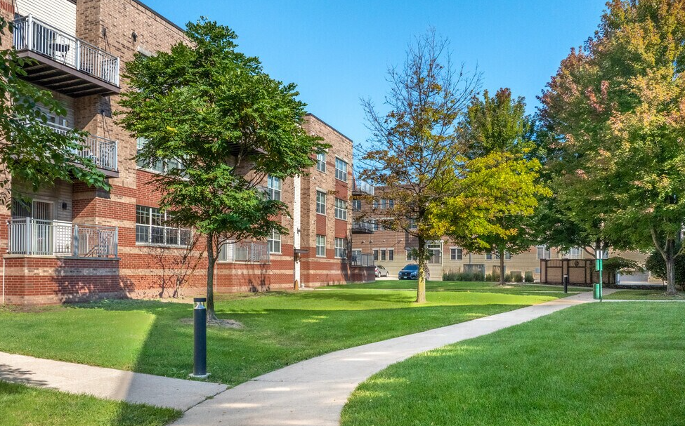 View of a green park with trees and a curved pathway, located next to a multi-story brick residential building on a sunny day.