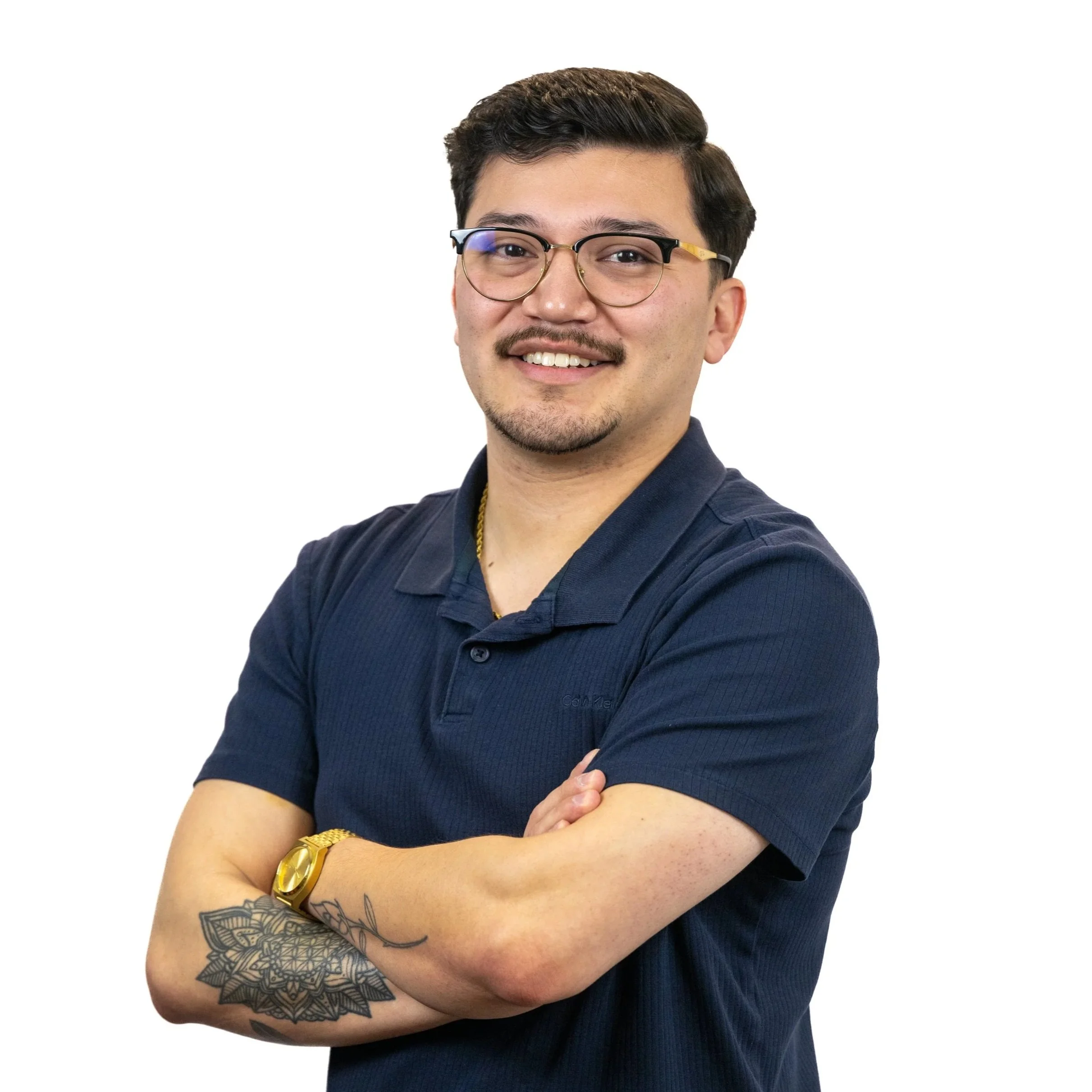 Portrait of a young man with glasses, a mustache, and beard, wearing a navy blue polo shirt, gold watch, and necklace, standing with arms crossed against a white background.