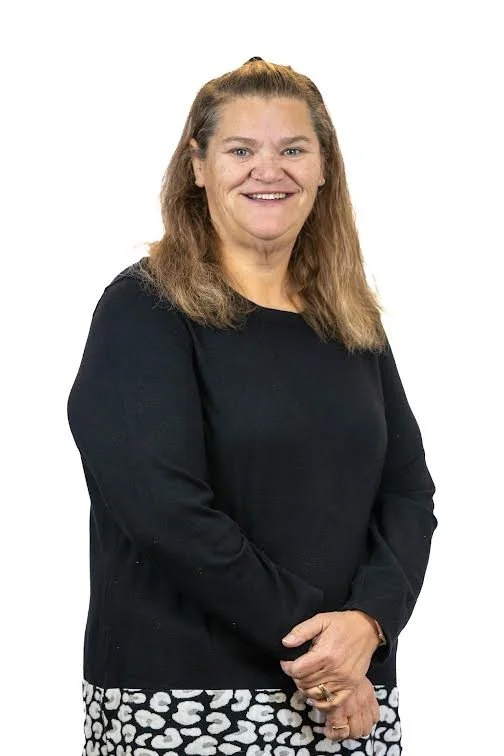 A smiling woman with shoulder-length brown hair, wearing a black sweater and a black-and-white patterned skirt, standing against a white background.